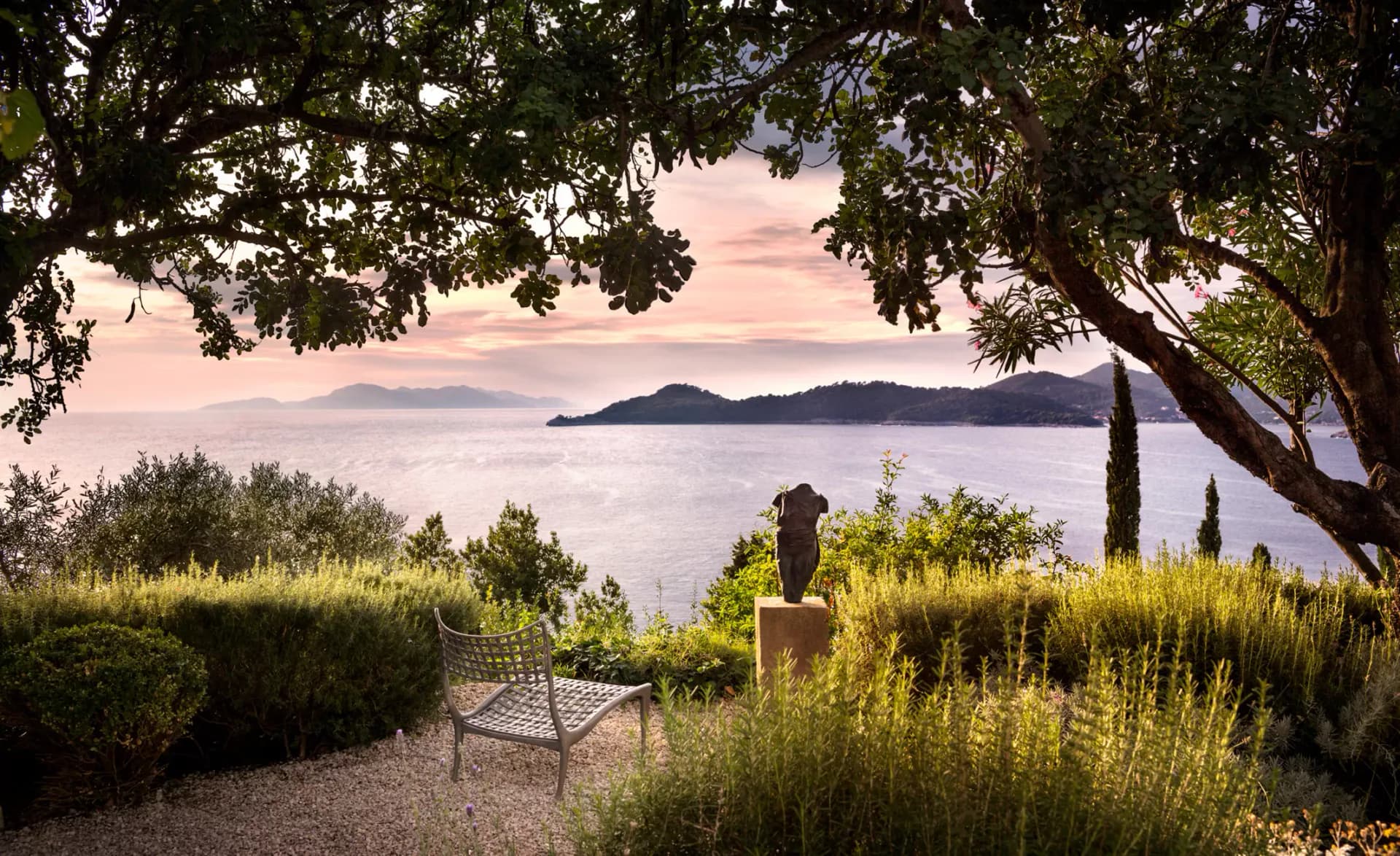 Coastal garden with Mediterranean plantings, wooden bench, and framed water view designed by Cate Steinacker.