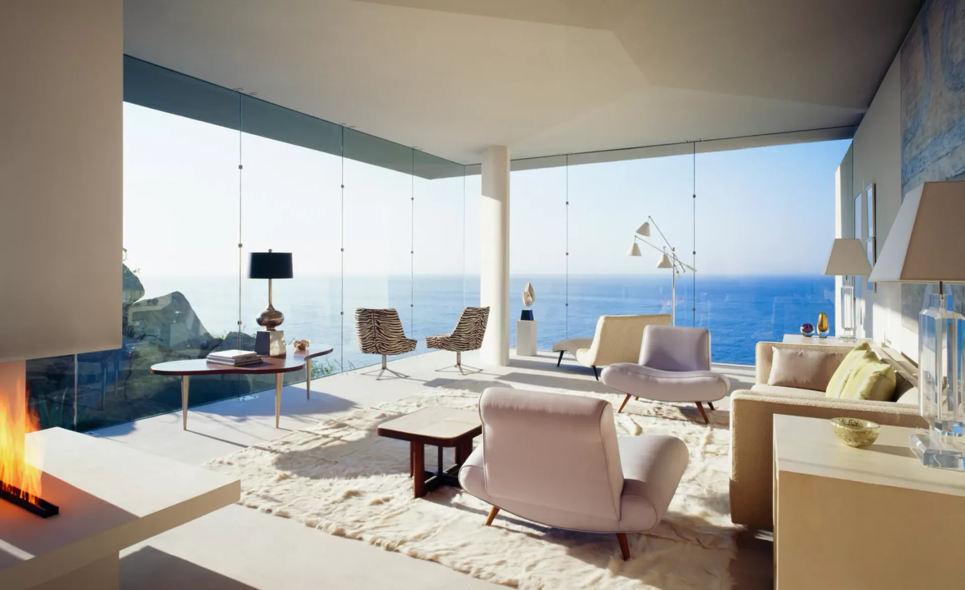 Luxury coastal living room with floor-to-ceiling sea views, featuring mid-century modern furniture, warm wood tones, and minimalist decor by Cate Steinacker.