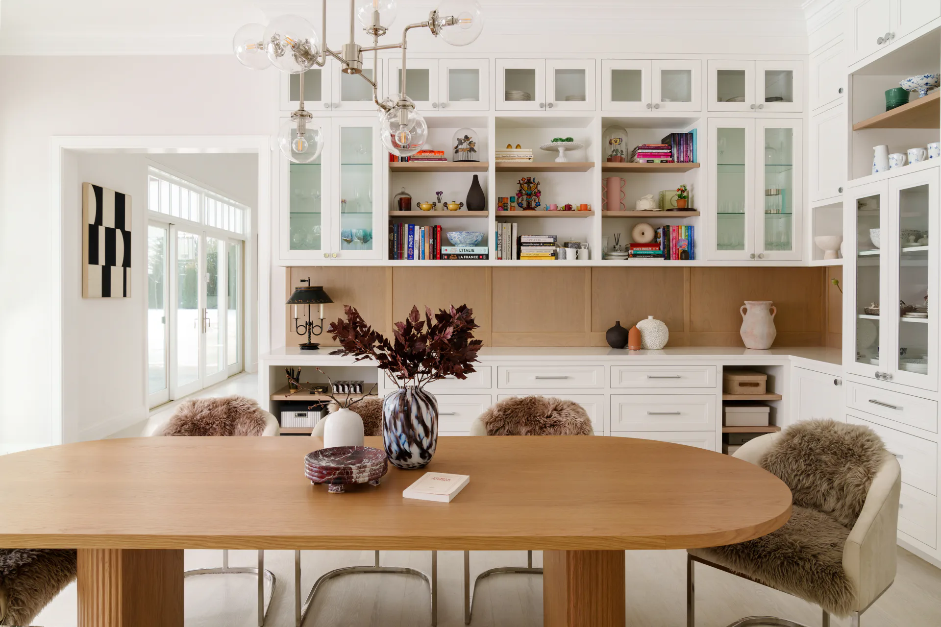 Bright modern kitchen and dining space with white cabinetry, glass-front upper cabinets, natural wood oval table, and layered shelving by Cecile Mattei Ekstrom.