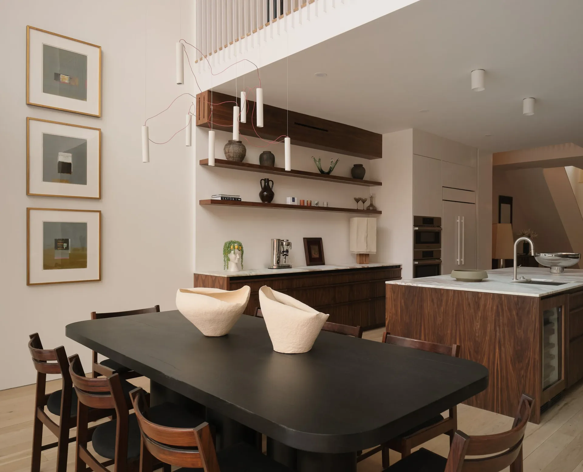 Modern open-plan kitchen and dining space with black dining table, dark wood cabinetry, floating shelves, and white pendant lighting, designed by Cem Evirgen.
