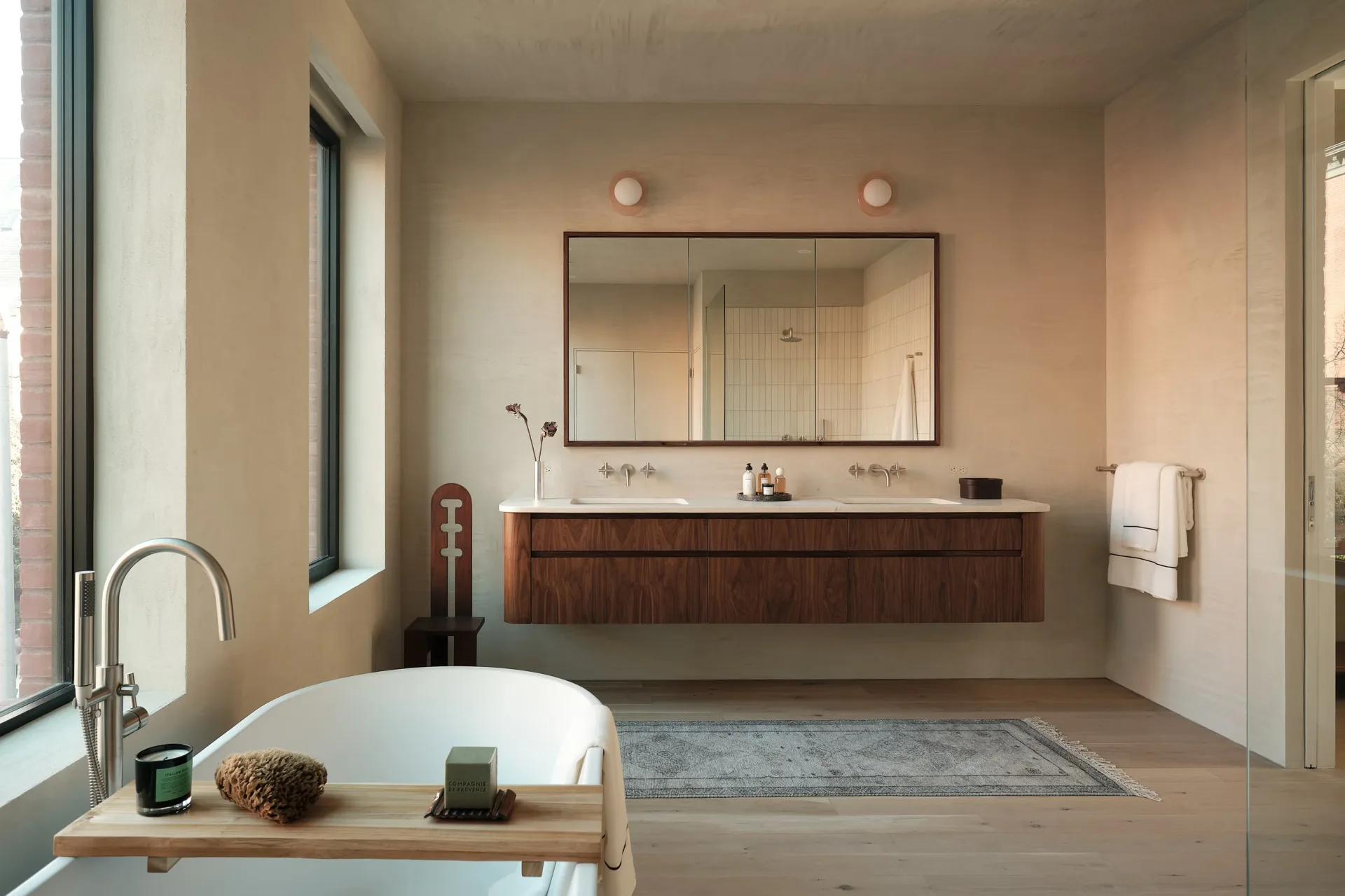 Minimalist bathroom with warm beige walls, freestanding white sofa tub, dark wood floating vanity, and large mirror by Cem Evirgen.