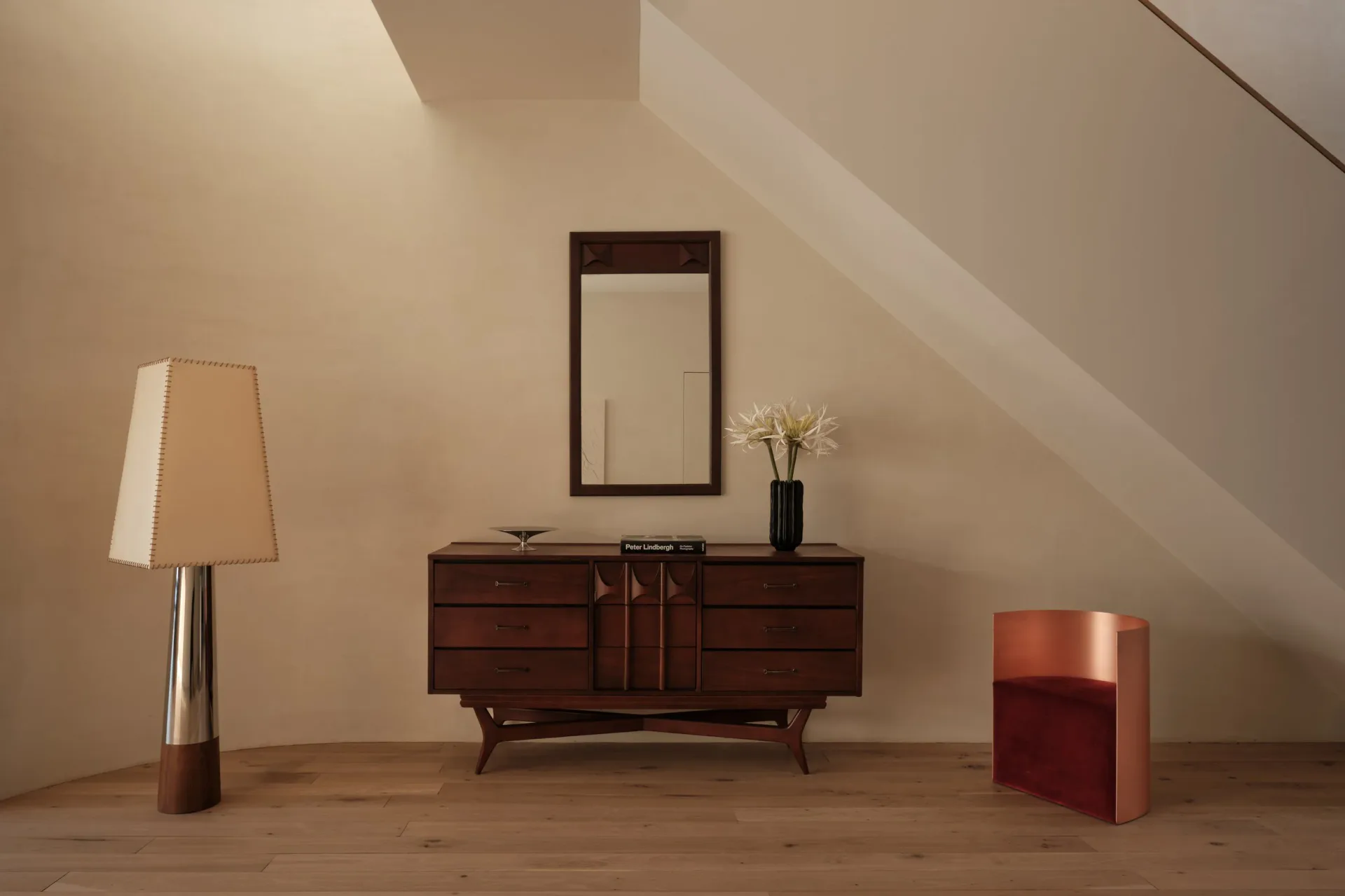 Minimalist mid-century modern interior with warm beige walls, dark wood credenza, framed artwork, floor lamp, potted flowers, and geometric red cube furniture by Cem Evirgen.