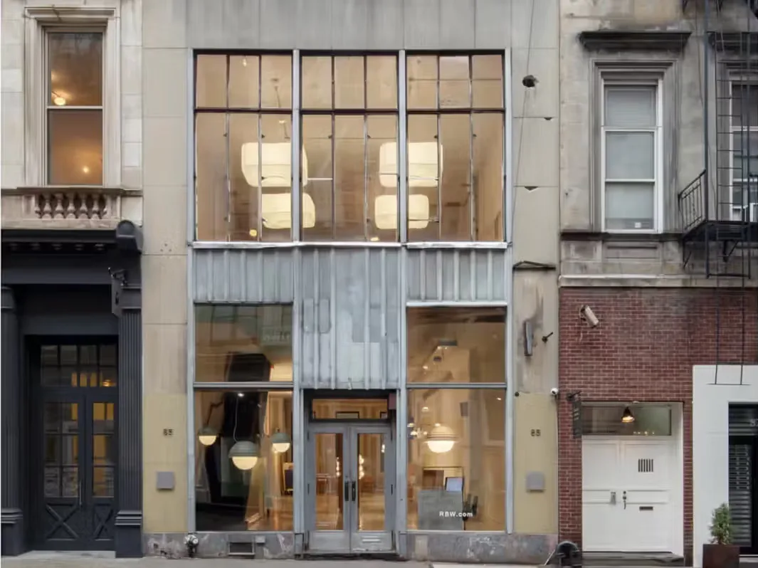 Storefront facade with large gridded windows, concrete and brick exterior, minimalist interior visible with warm lighting and pendant fixtures by Charles Brill.