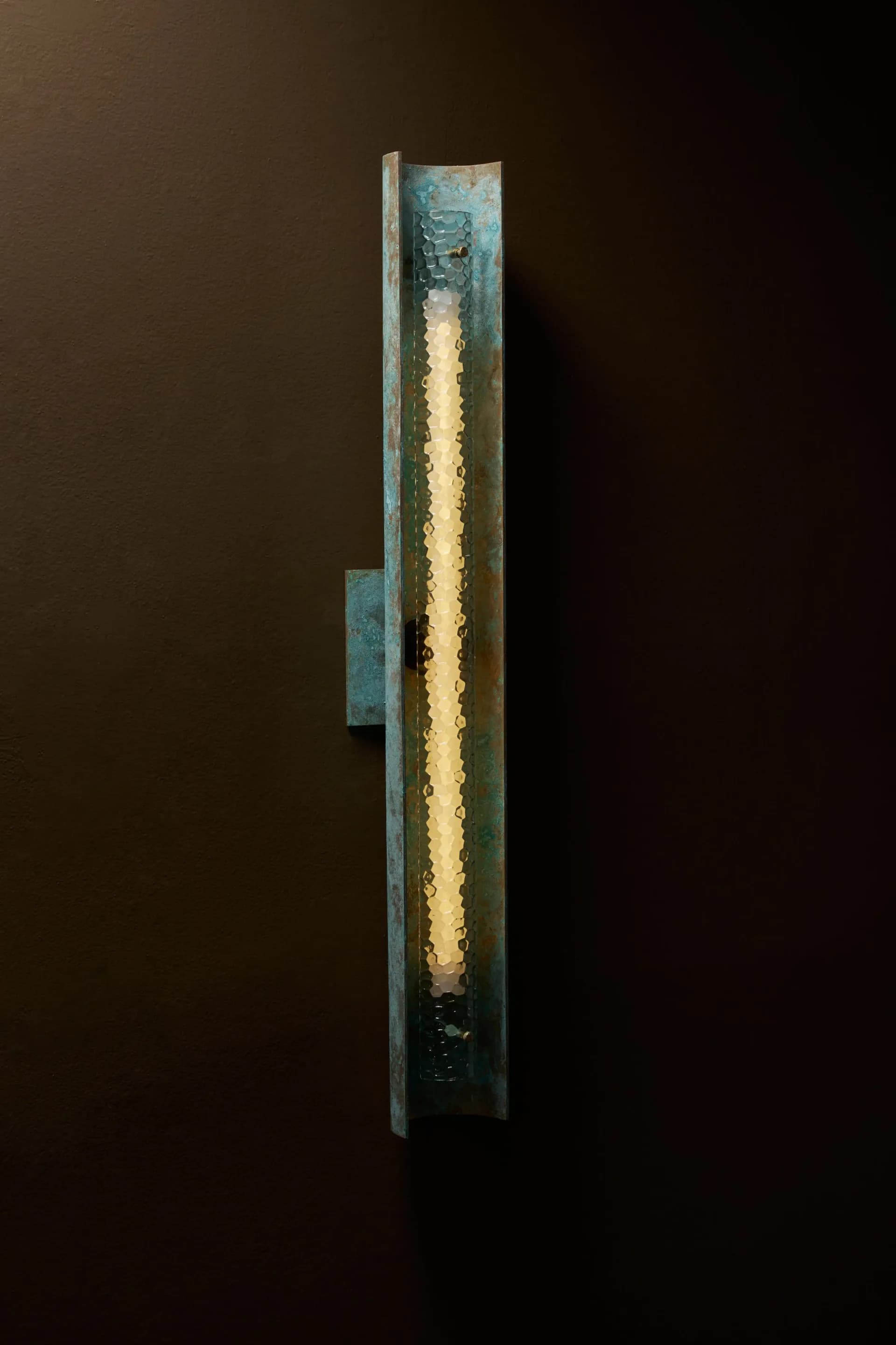 Tall vertical wall-mounted lighting installation with weathered patina metal frame and glowing warm-toned linear element by Charles Grantham.