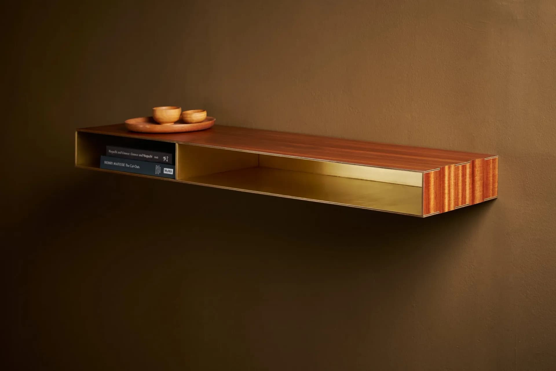 Golden-bronze floating shelf with ribbed copper detailing, small sculptural objects on top, and dark cabinetry beneath, set against warm brown background by Charles Grantham.