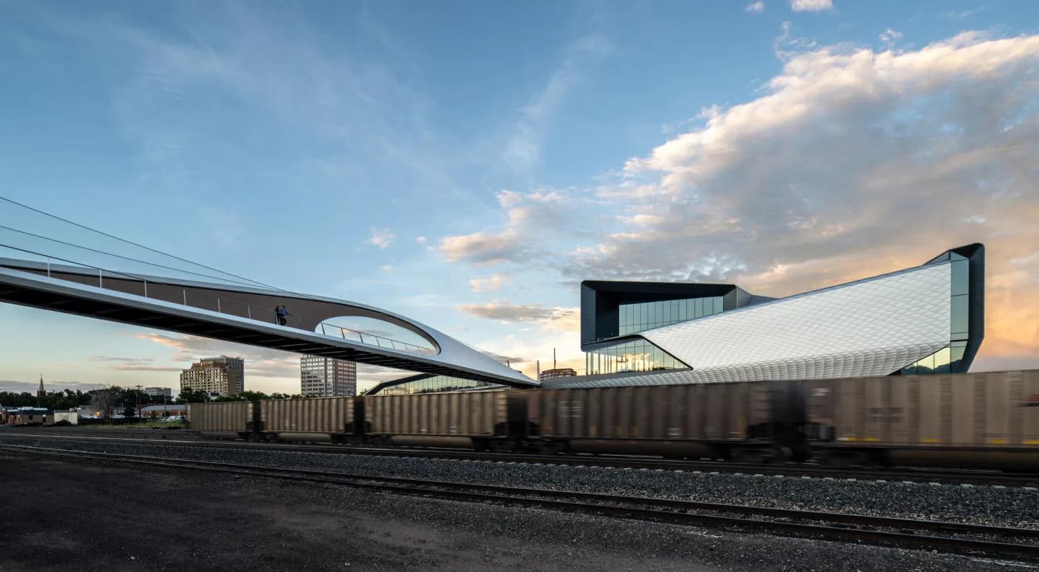 Modern architectural structure with sweeping curved canopy suspended over railroad tracks against dramatic sky by Charles Renfro.