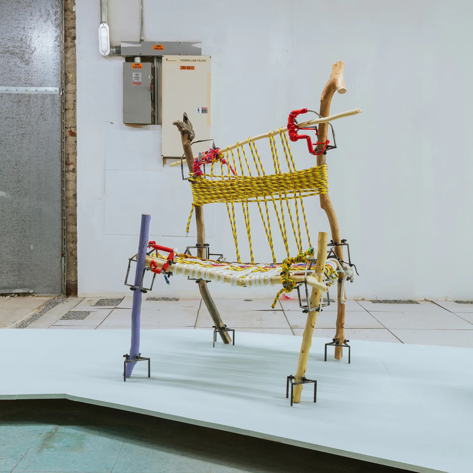 Sculptural installation featuring stacked wooden furniture frames wrapped in yellow rope with red metal connectors, displayed in a gallery space by Chen Chen.