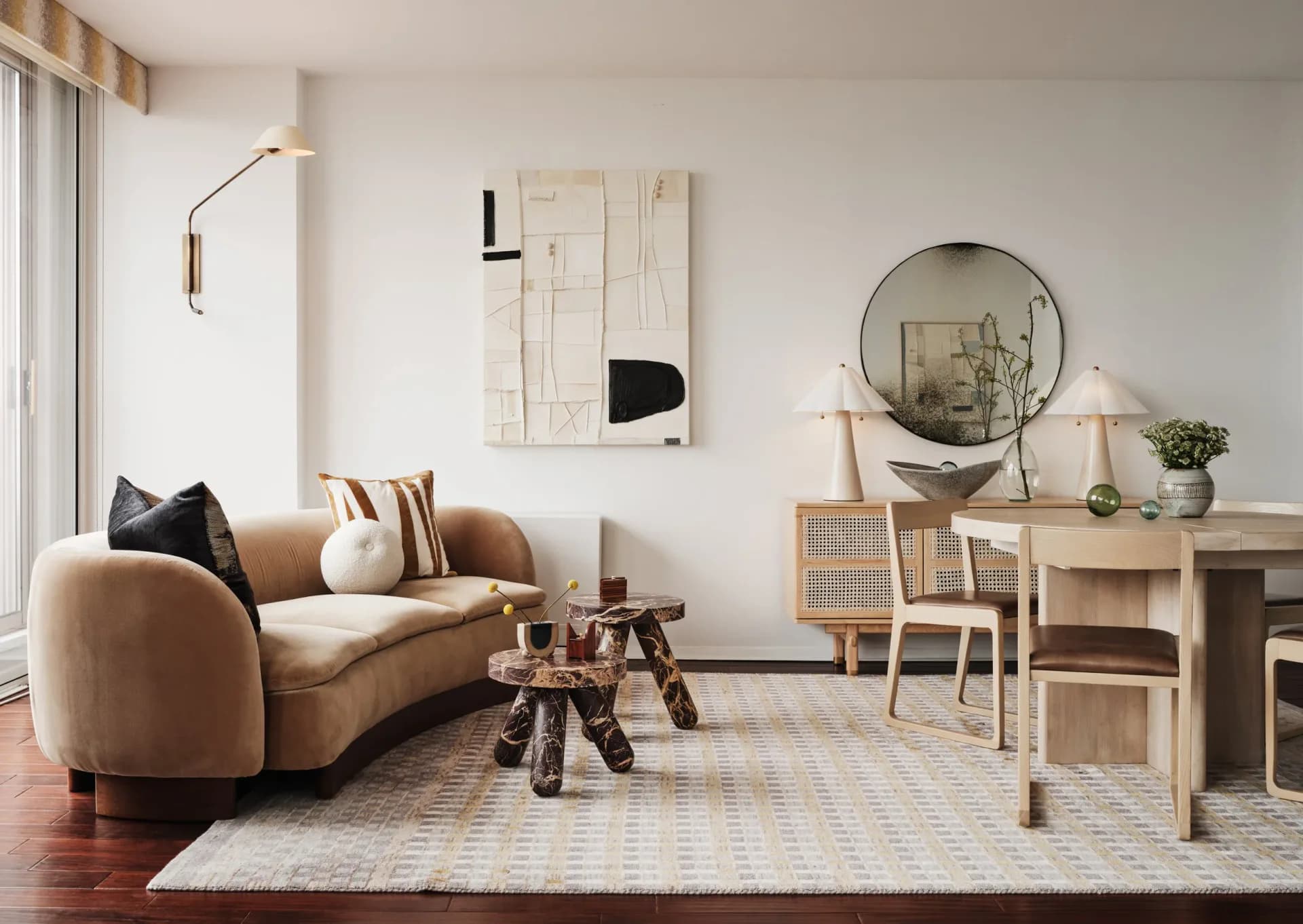 Light-filled living room interior with curved tan sofa, wooden desk, round mirror, and neutral palette by Cherie Urrutia.