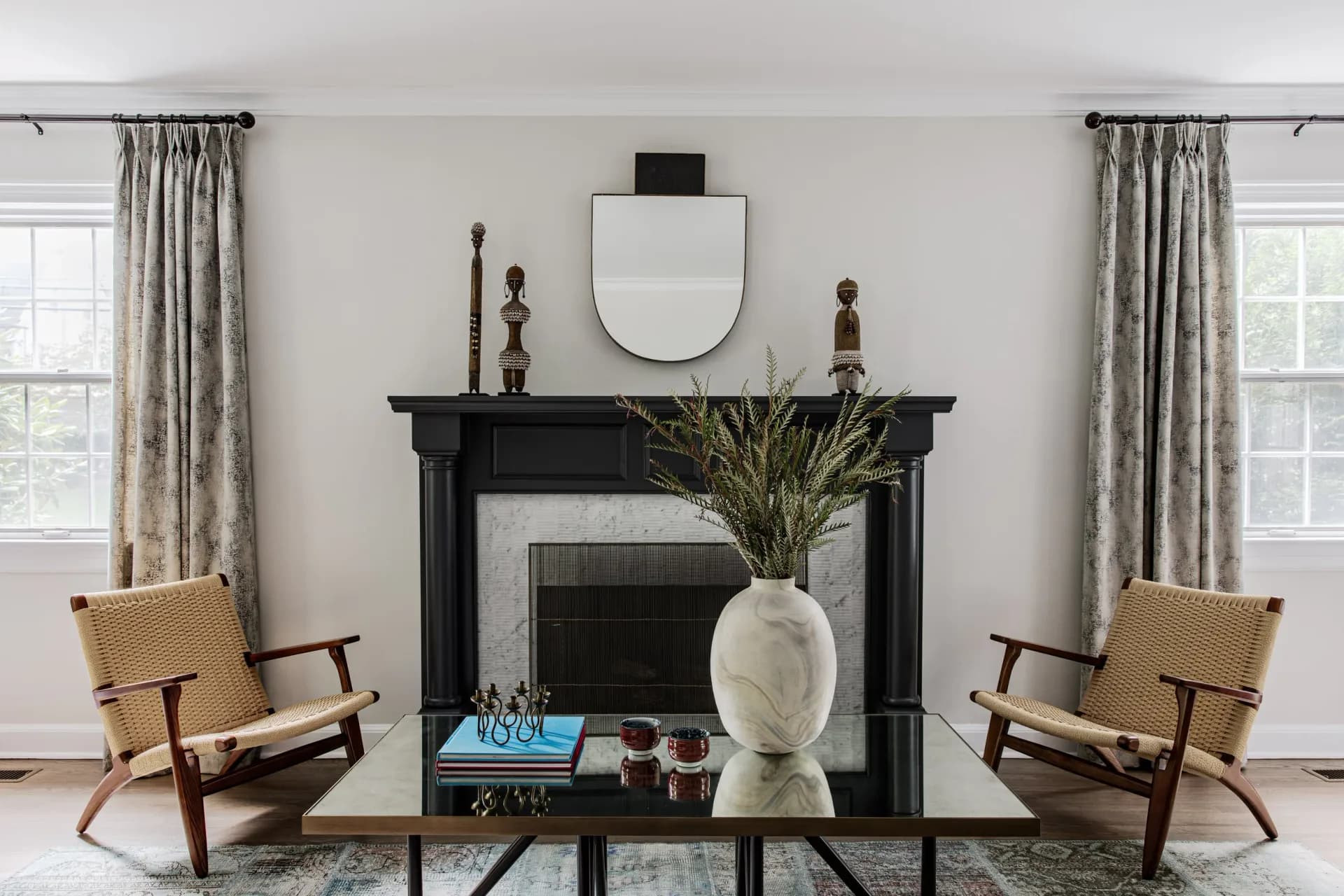 A symmetrical living room features a black fireplace mantel centered between two wood-frame chairs, flanked by tall windows with striped curtains, in a neutral palette with brass accents, designed by Cherie Urrutia.
