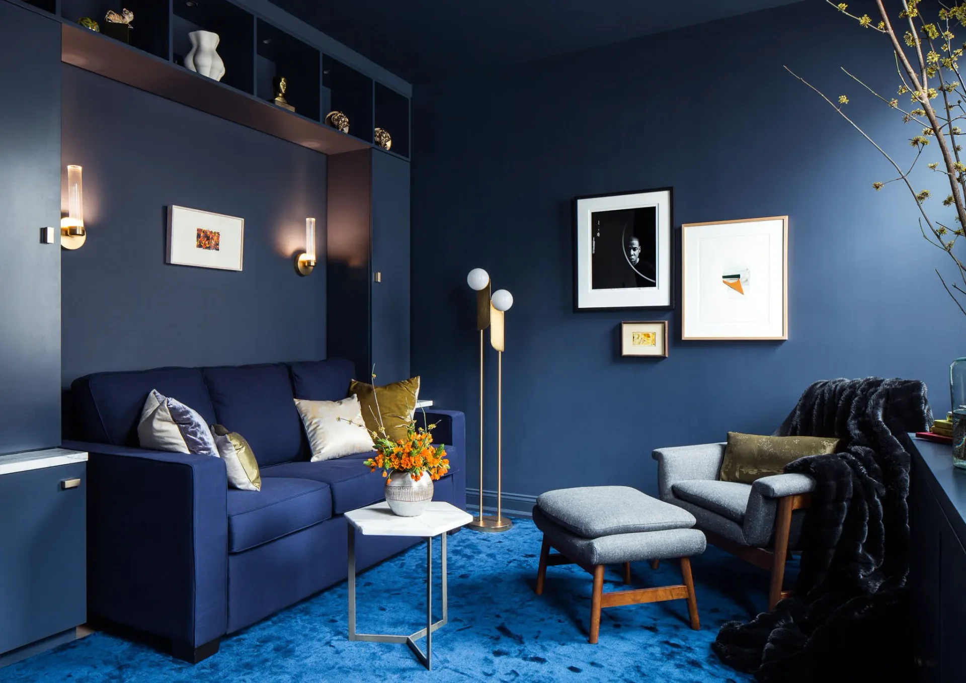 Modern living room with deep blue walls, navy velvet sofa, light gray armchair, glass coffee table, and curated gallery wall featuring framed artwork and photographs by Cherie Urrutia.
