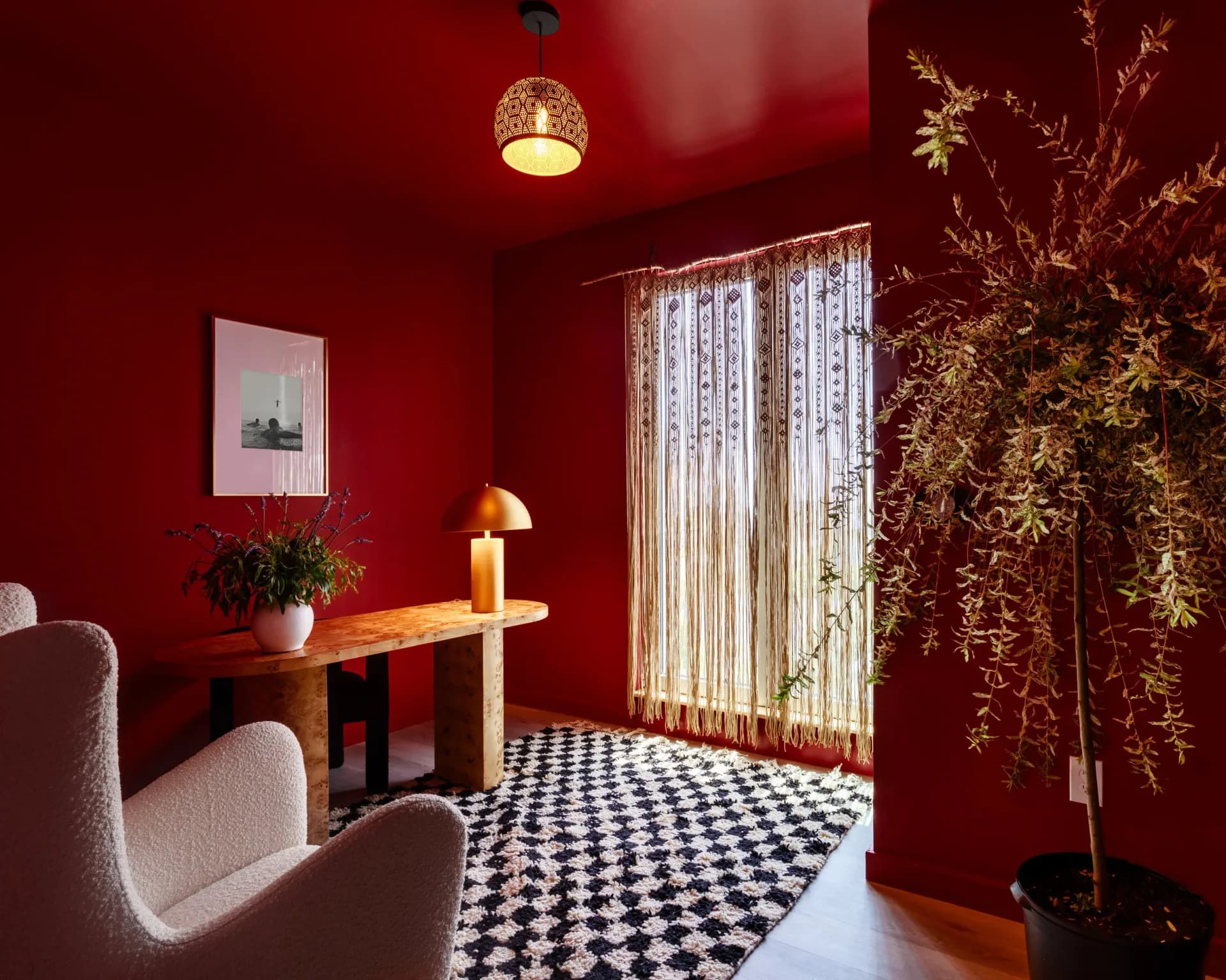 Bold red interior room with checkered floor, cream-colored chair, wooden desk, pendant lighting, and golden dried plant installation by Cherub Stewart.