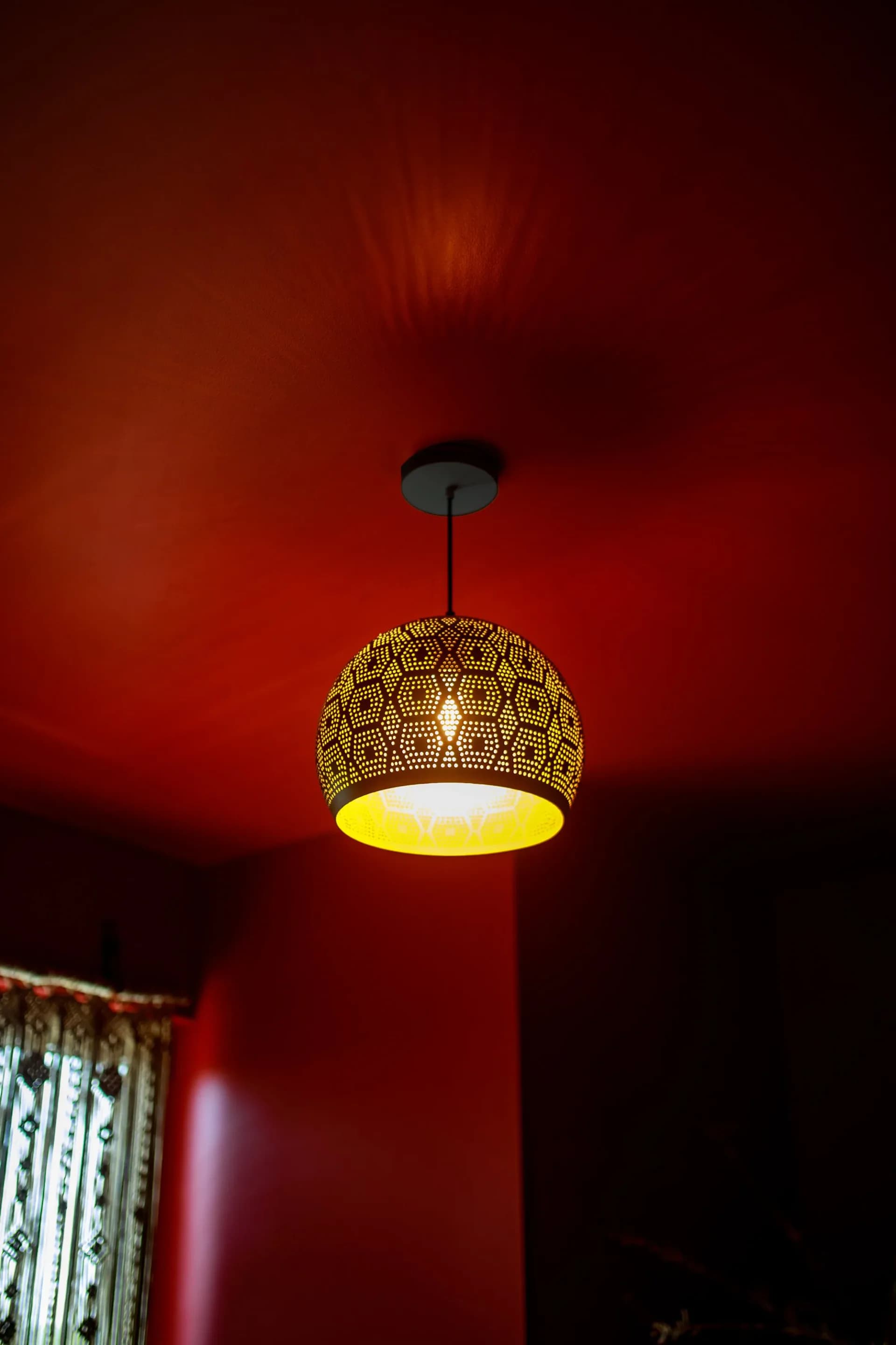 Pendant lighting fixture with perforated brass dome shade suspended against deep red wall, warm glow emanating from interior by Cherub Stewart.