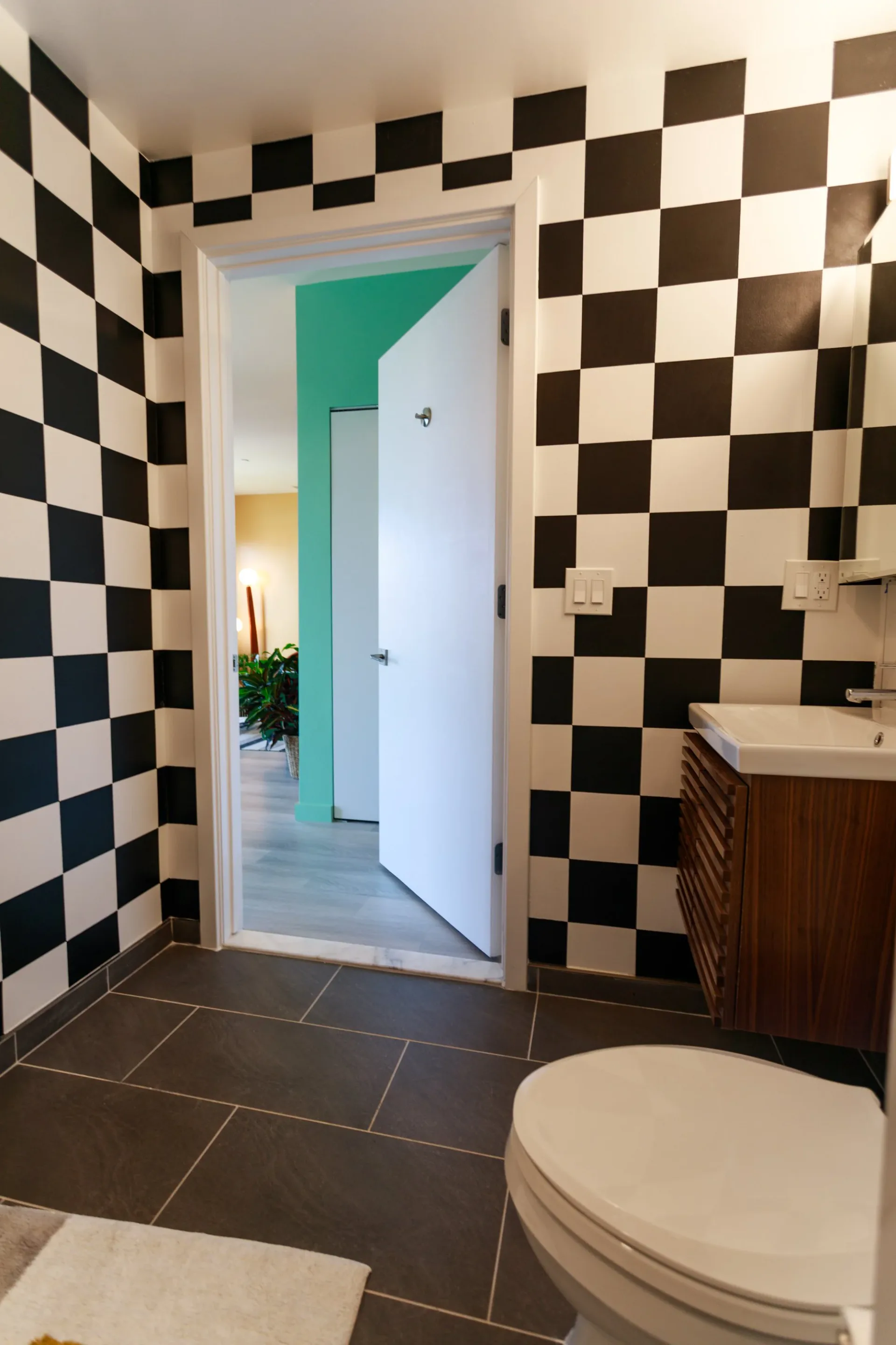 Black-and-white checkered bathroom with dark tile flooring, white toilet, wooden vanity, and open doorway revealing turquoise accent wall by Cherub Stewart.