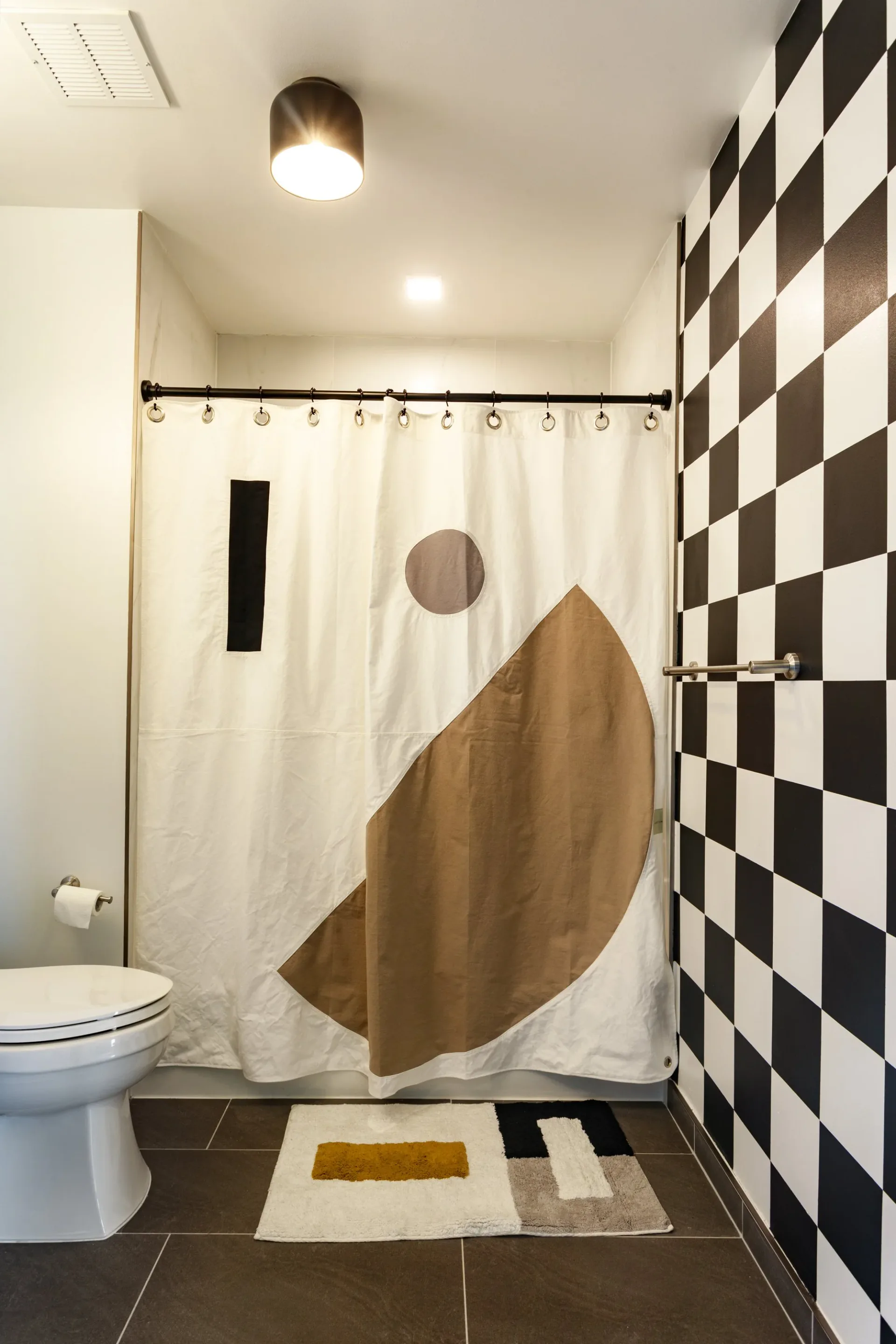 Modern bathroom with black and white checkered tile wall, cream shower curtain featuring abstract brown design, dark flooring, and geometric bath mat by Cherub Stewart.