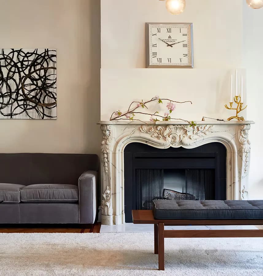 Elegant living room featuring a carved cream fireplace, dark upholstered sofa, wood coffee table, and gallery wall with abstract black artwork, designed by Cheryl Chase-Mackenna.