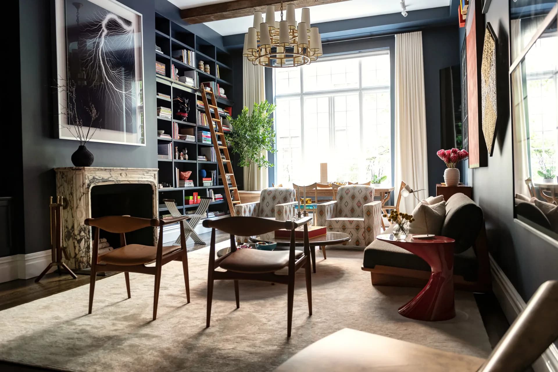 Contemporary living room with dark walls, built-in shelving, fireplace, mid-century furniture, brass chandelier, and large windows by Chrissy Gamba.