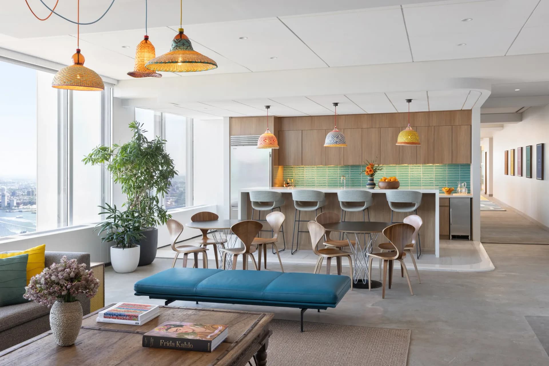 Modern open-plan office kitchen with wood cabinetry, green tile backsplash, pendant lighting, and lounge seating with blue bench by Chrissy Gamba.