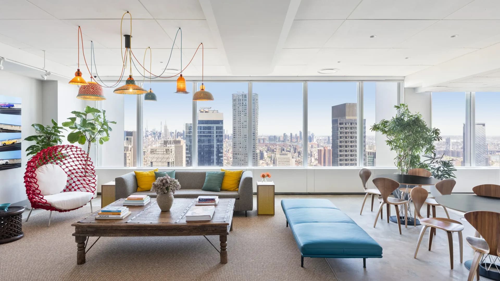 Modern high-rise living room with floor-to-ceiling NYC skyline views, gray sectional with yellow accents, brass pendant lights, blue bench seating, and potted plants by Chrissy Gamba.