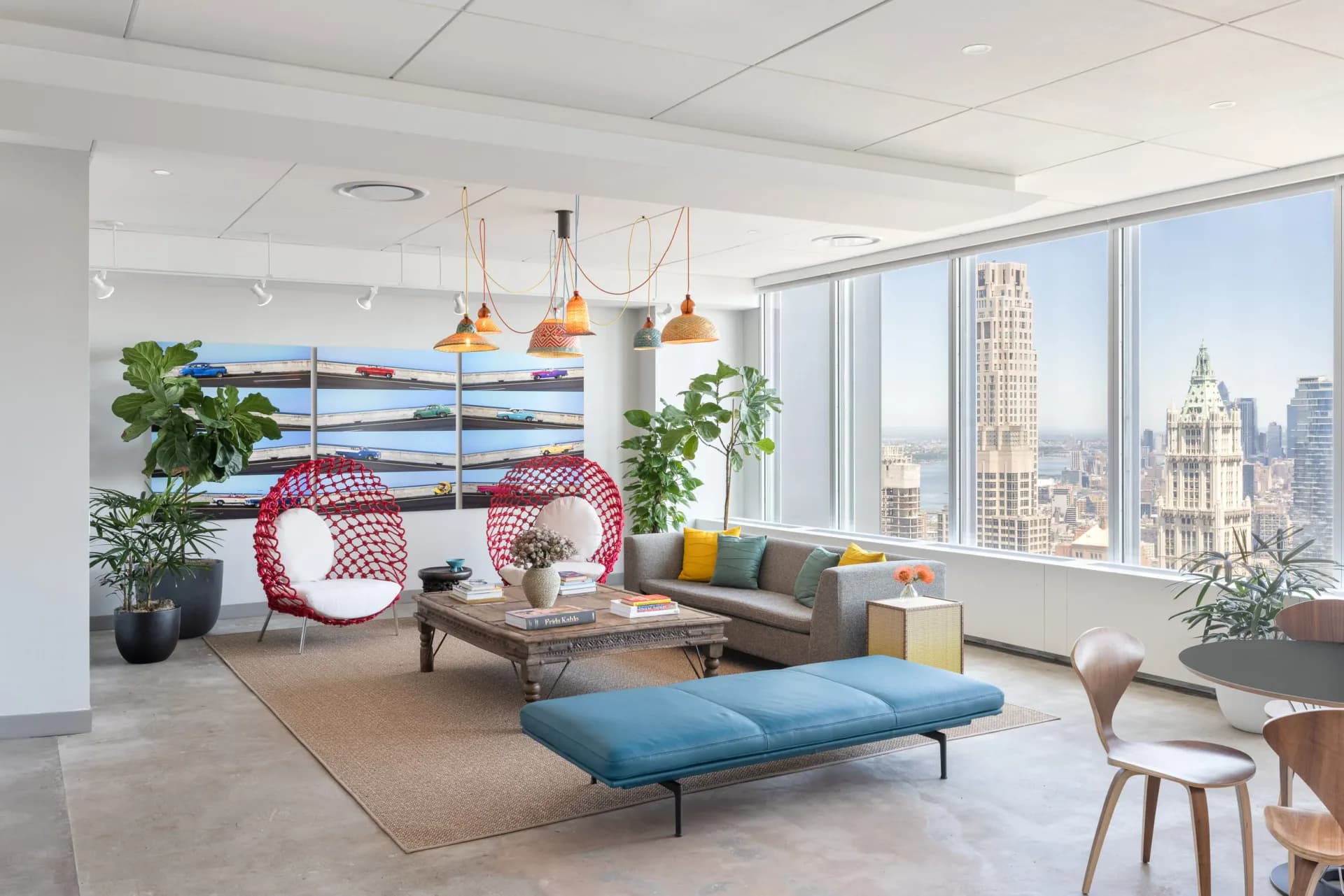 Modern high-rise office lounge with city skyline views, featuring copper pendant lights, red accent chair, blue bench seating, potted plants, and neutral palette by Chrissy Gamba.