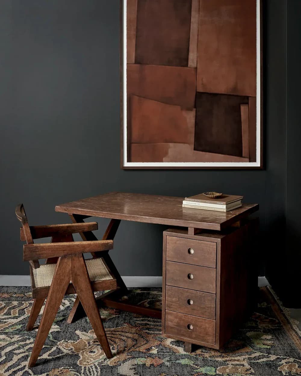 Modern workspace with walnut desk and matching chair featuring woven seat, styled with books and artwork, against charcoal wall by Christiane Lemieux.