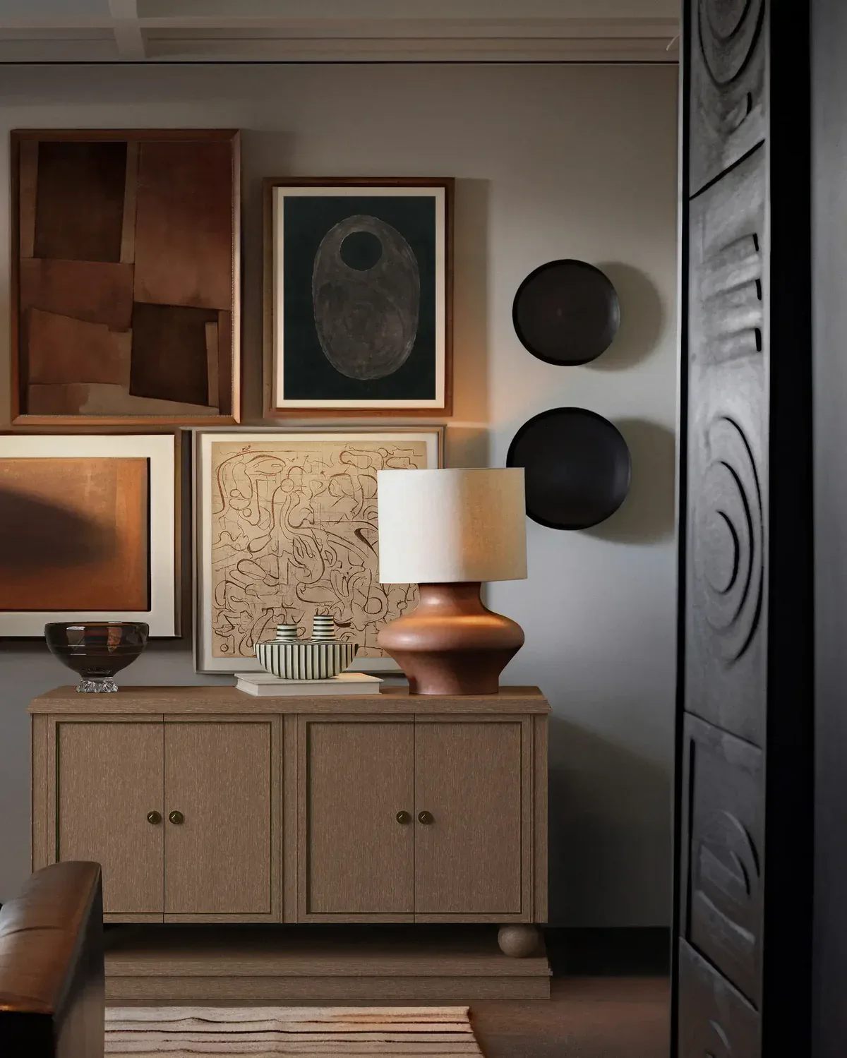 Warm-toned entryway with light wood cabinetry, copper table lamp, curated art gallery wall, and black sculptural spheres by Christiane Lemieux.