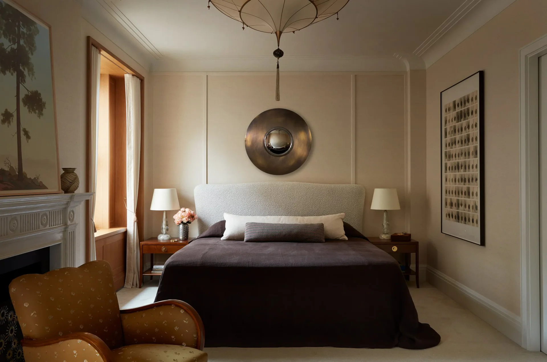 Serene bedroom with dark bed, cream walls, brass circular wall sculpture, warm table lamps, and caramel leather chair by Christine Gachot.