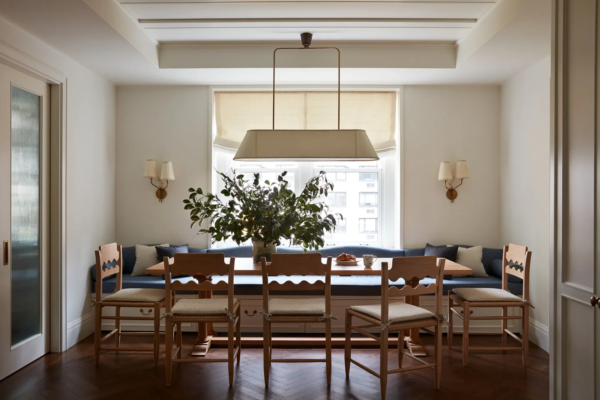 Dining room with wooden table and chairs, blue upholstered seats, brass pendant light, potted plant centerpiece, and cream walls by Christine Gachot.