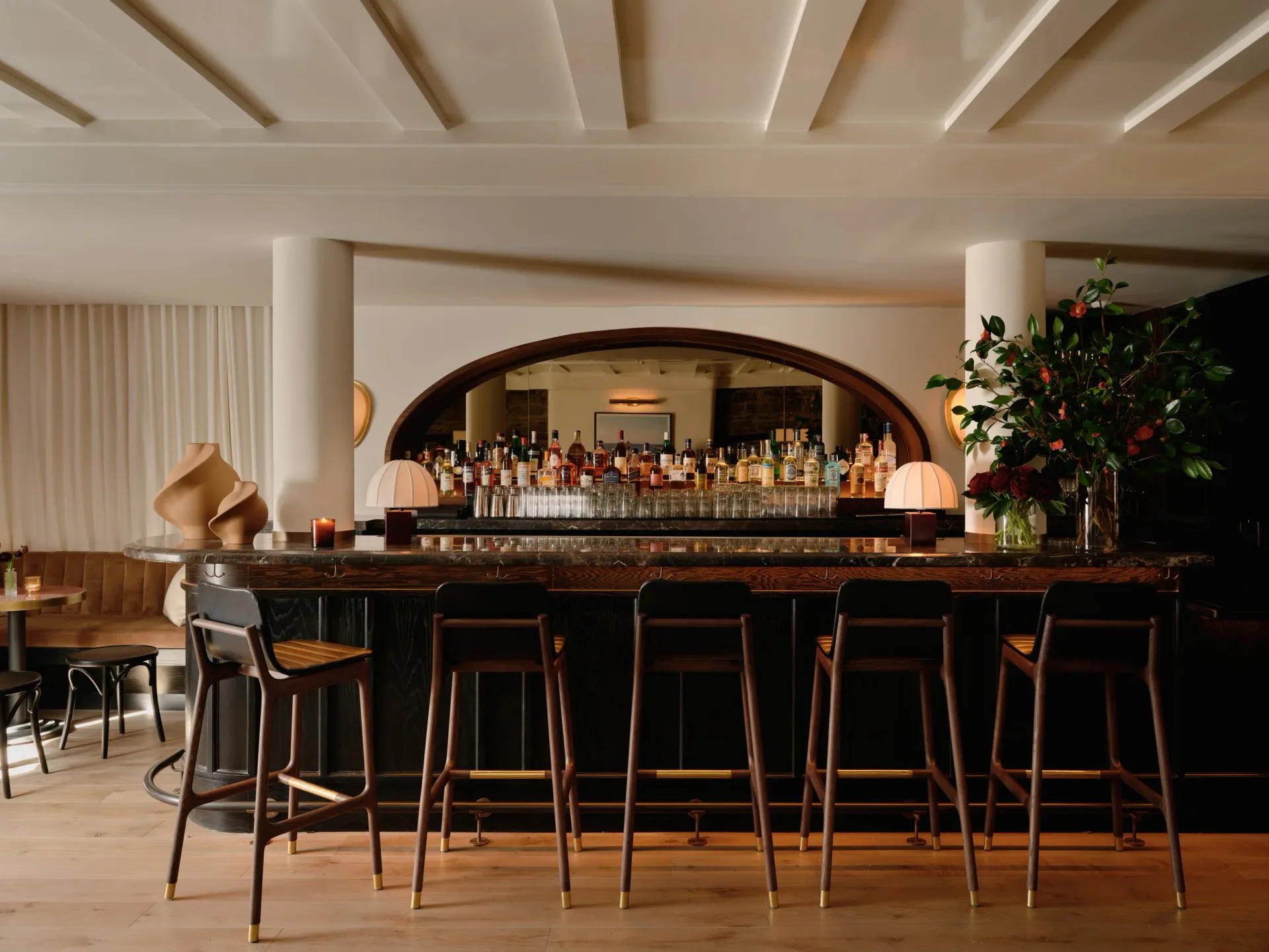 Elegant bar interior with curved archway, wooden bar stools, dark wood counter, and shelved bottles, designed by Christine Gachot.