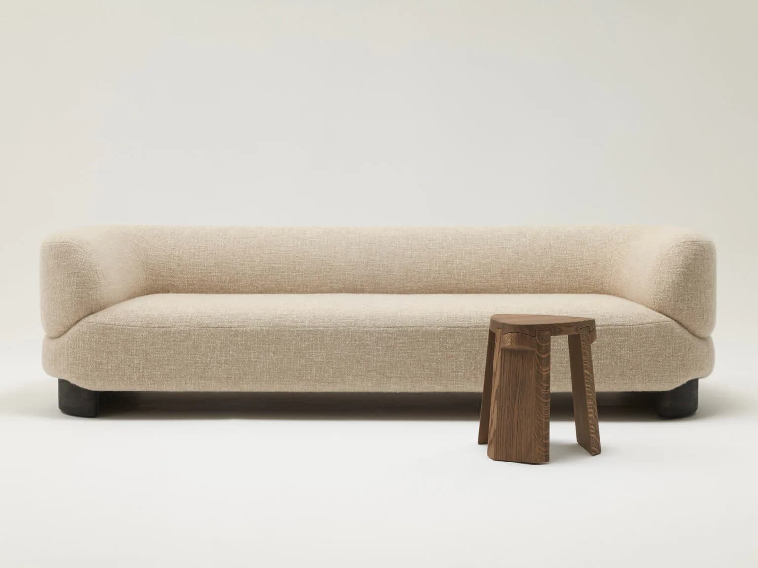 Minimalist cream linen sofa with rounded arms and black base, paired with wooden stools, against neutral background by Christophe Delcourt.