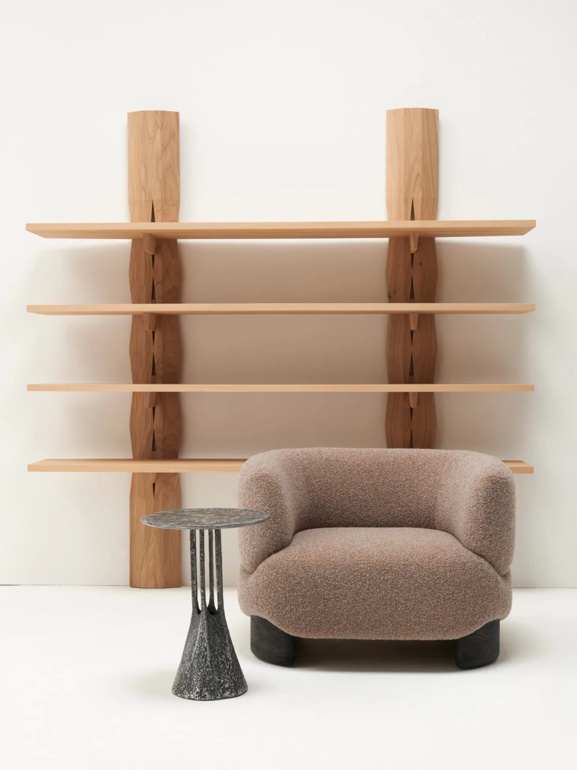 Wooden wall-mounted shelving unit with vertical posts and horizontal shelves in light wood, paired with a curved upholstered armchair in beige fabric and a modernist side table by Christophe Delcourt.