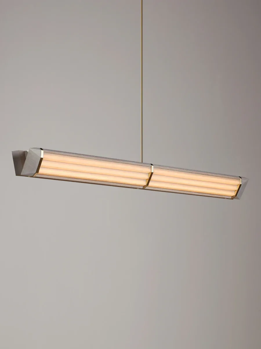 Suspended linear pendant light fixture with elongated cylindrical glass diffusers and brushed metal housing, emitting warm light against neutral background, by Christopher Beardsley.