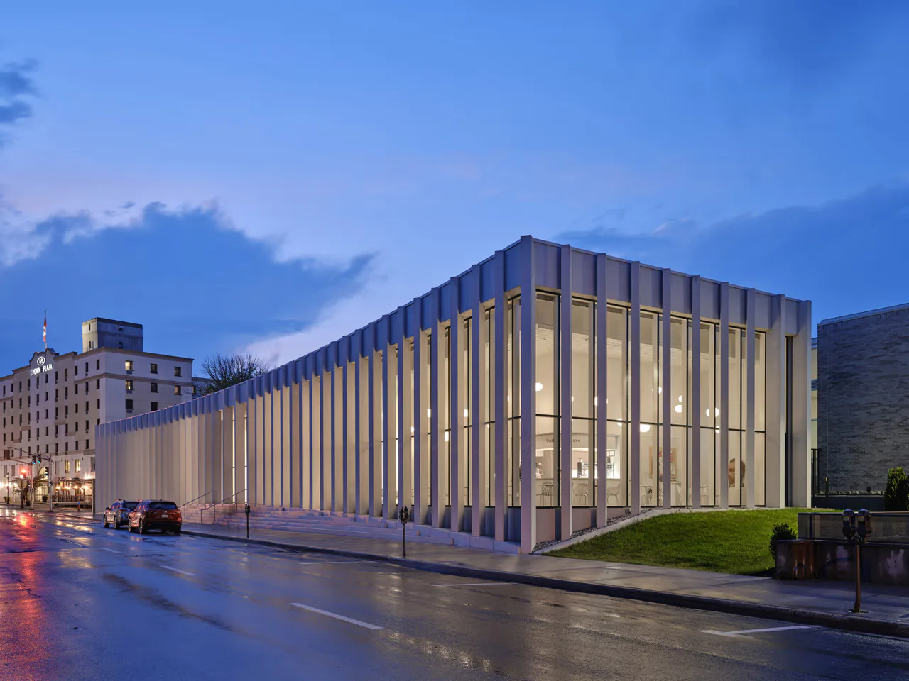 Modern institutional building with illuminated vertical metal-clad facade at dusk, featuring clean geometric lines and glass panels, wet street foreground by Christopher Cheap.