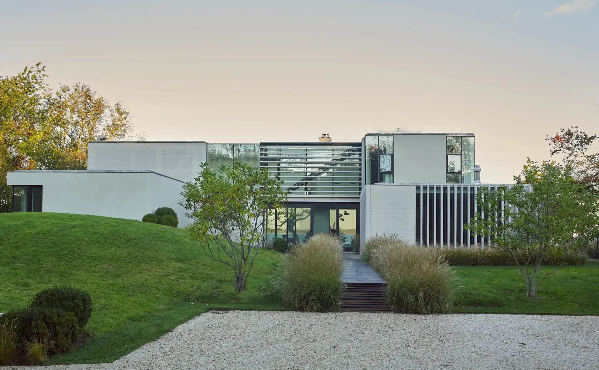 Modern residential building with white and dark metal cladding, extensive glazing, and wooden deck set on manicured green landscape with gravel pathways by Christopher Coy.