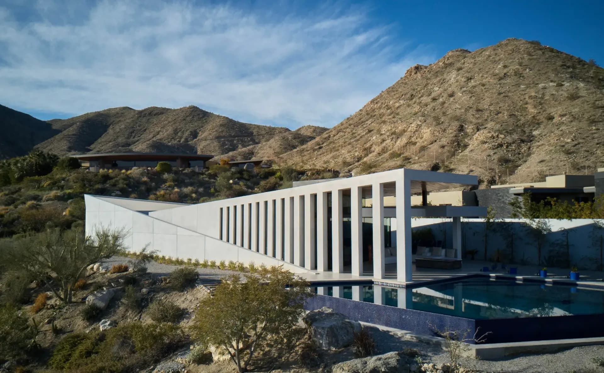 Contemporary residential architecture with white vertical louvers overlooking a desert landscape with mountains, featuring a reflecting pool by Christopher Coy.