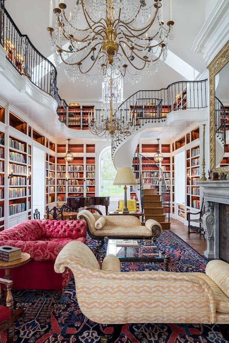 A two-story library room features floor-to-ceiling bookshelves, a crystal chandelier, pink velvet seating, patterned upholstered chaise lounges, and a fireplace by Christopher Labrot.