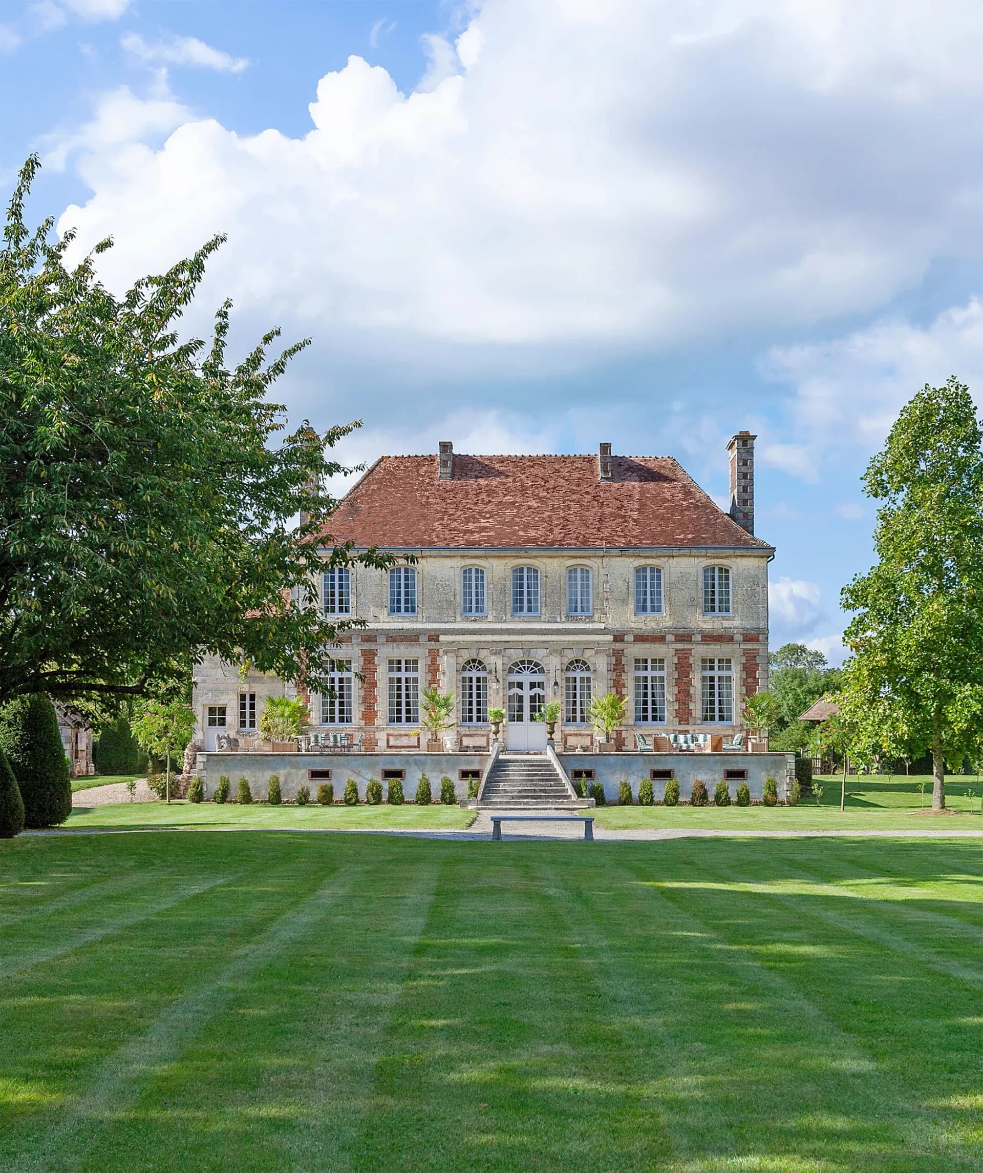 Neoclassical château with terracotta roof and cream-colored façade faces expansive manicured lawn with central bench, framed by mature trees by Christopher Labrot.