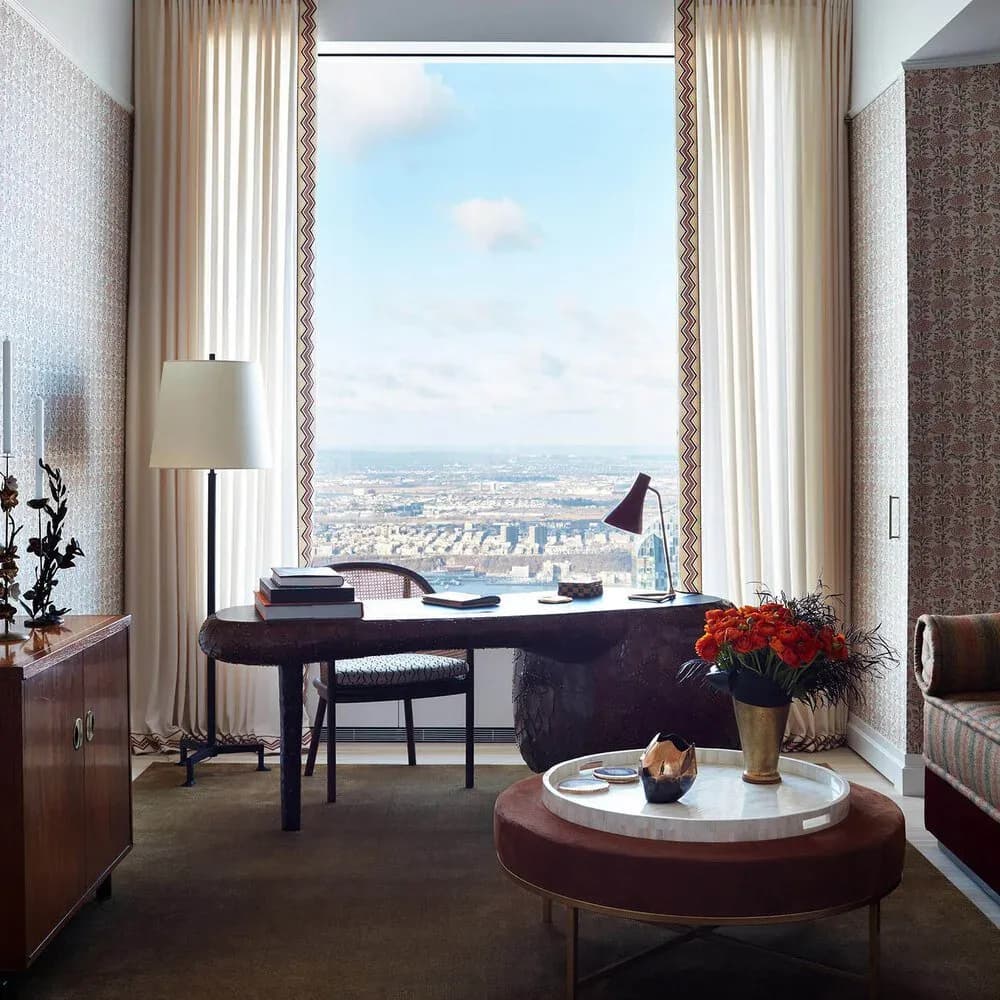 High-rise penthouse office with floor-to-ceiling windows overlooking a cityscape, dark wood desk, round marble side table, and neutral drapery by Christopher Labrot.
