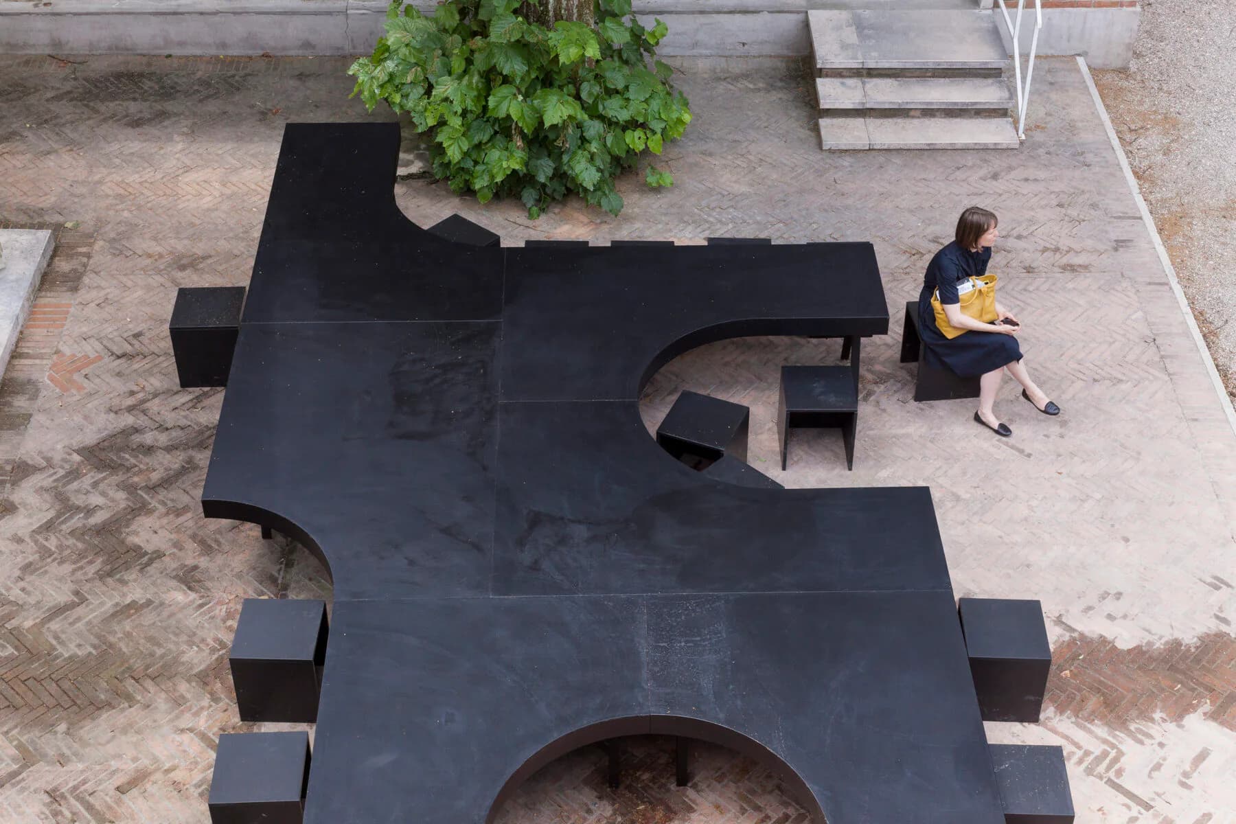 Overhead view of a black sculptural installation in a courtyard with geometric angular forms and a circular opening, designed by Christopher Leong.