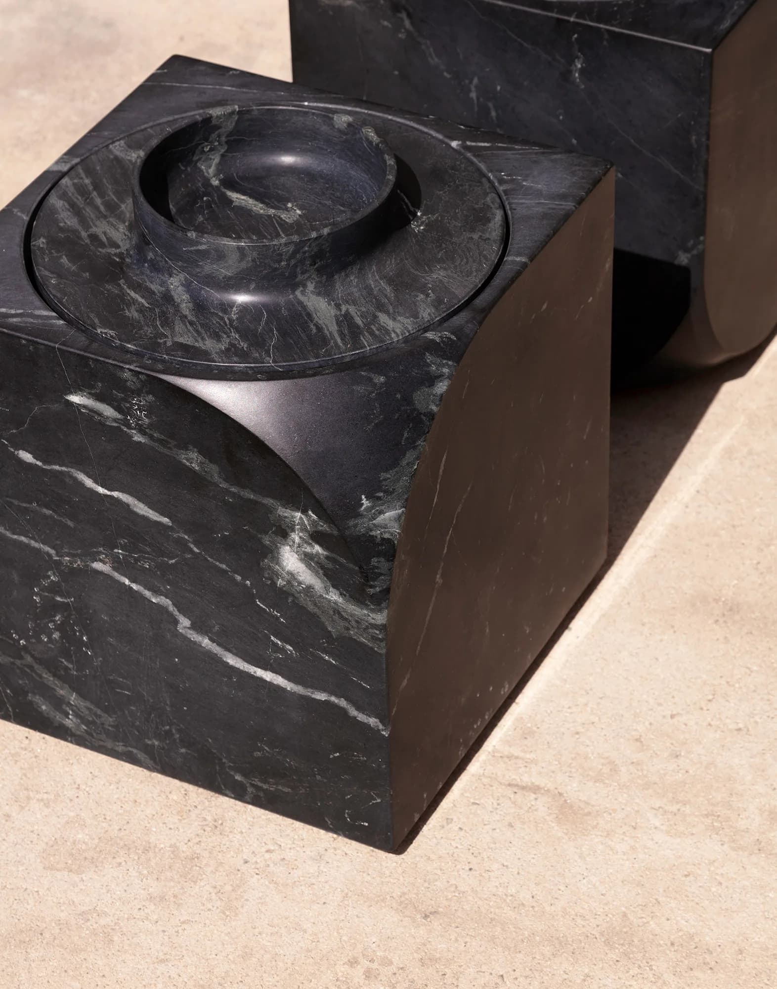 Black marble sculptural furniture piece with circular bowl-like top element and geometric block form, photographed outdoors by Christopher Leong.