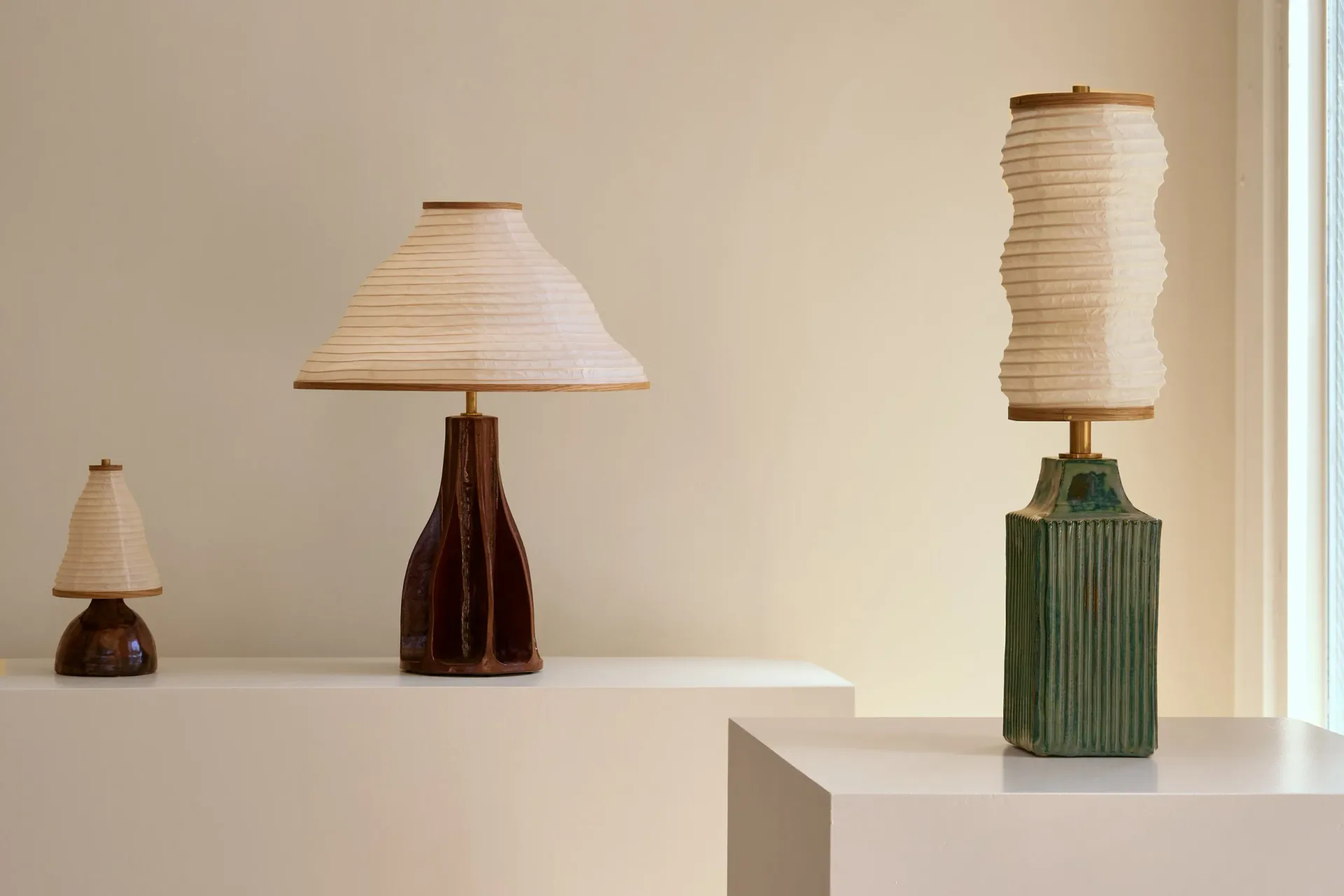 Three ceramic table lamps with paper shades in varying heights displayed on white platforms, featuring dark and green bases, designed by Christopher Merchant.