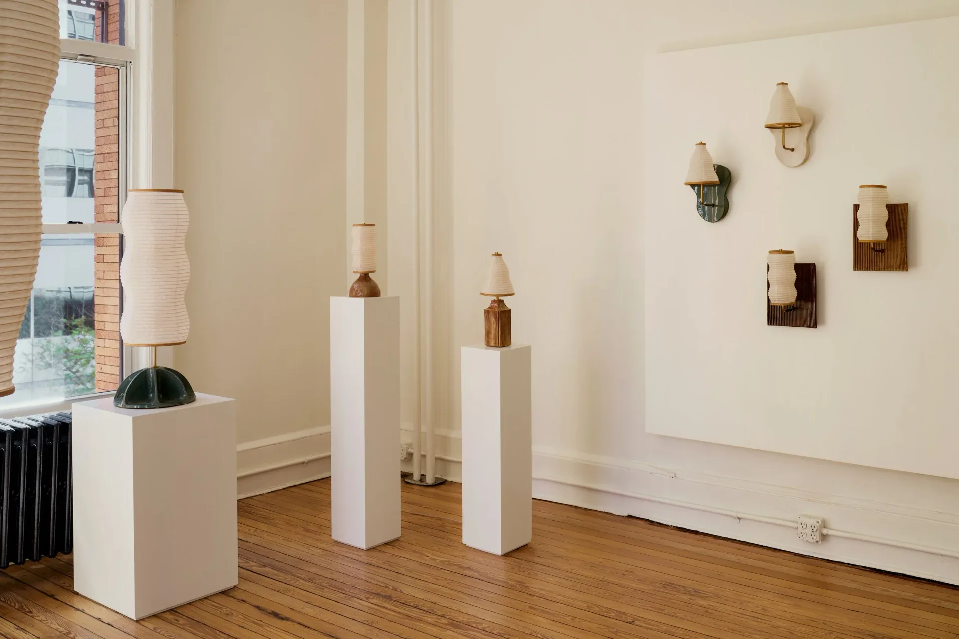 Gallery installation with white pedestals displaying amber glass vessels and wall-mounted wooden sculptural pieces against cream walls and wood flooring by Christopher Merchant.
