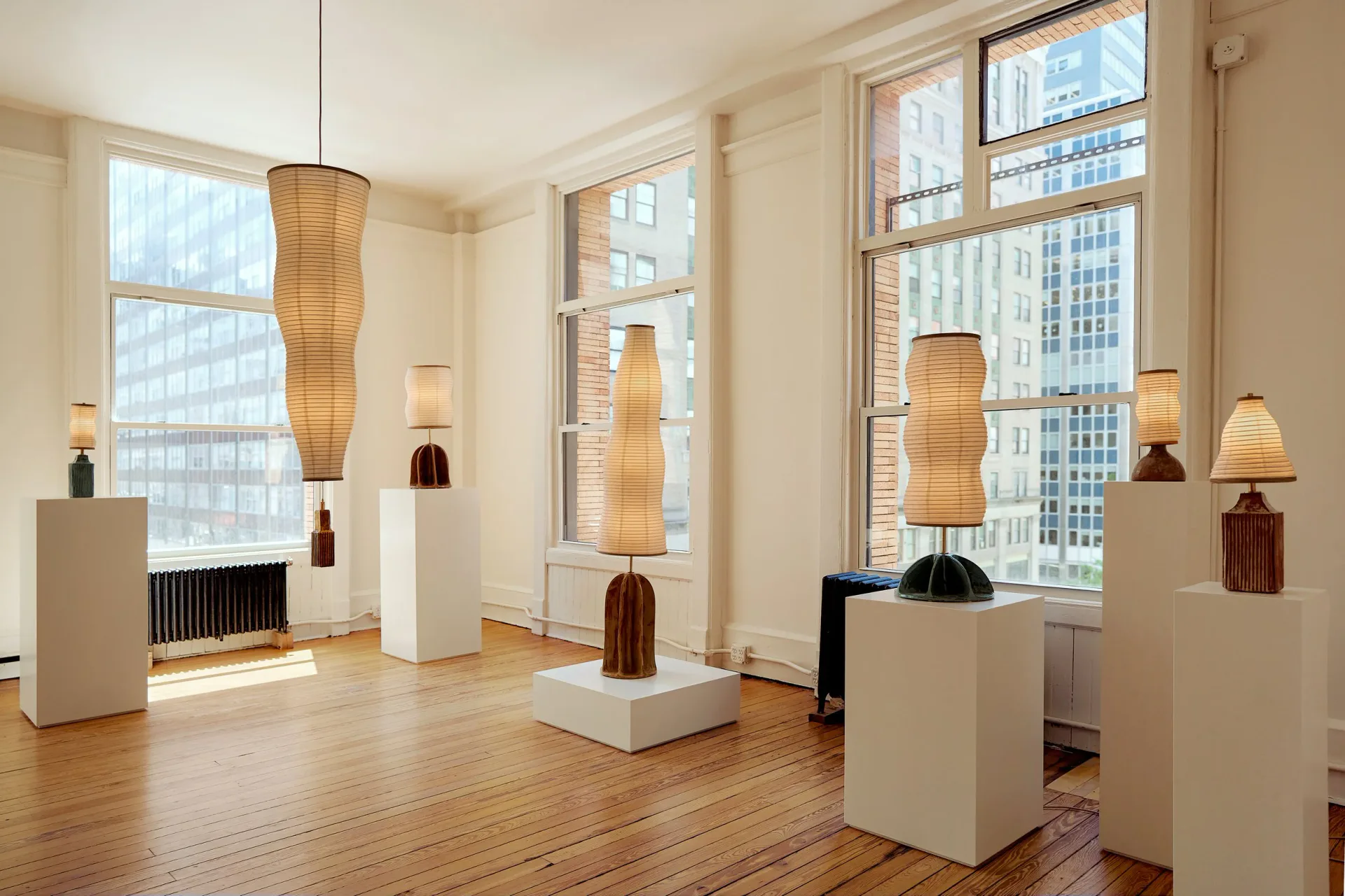 Bright gallery space with tall windows, woven pendant lights, sculptural vessels on pedestals, warm wood flooring, and city views by Christopher Merchant.