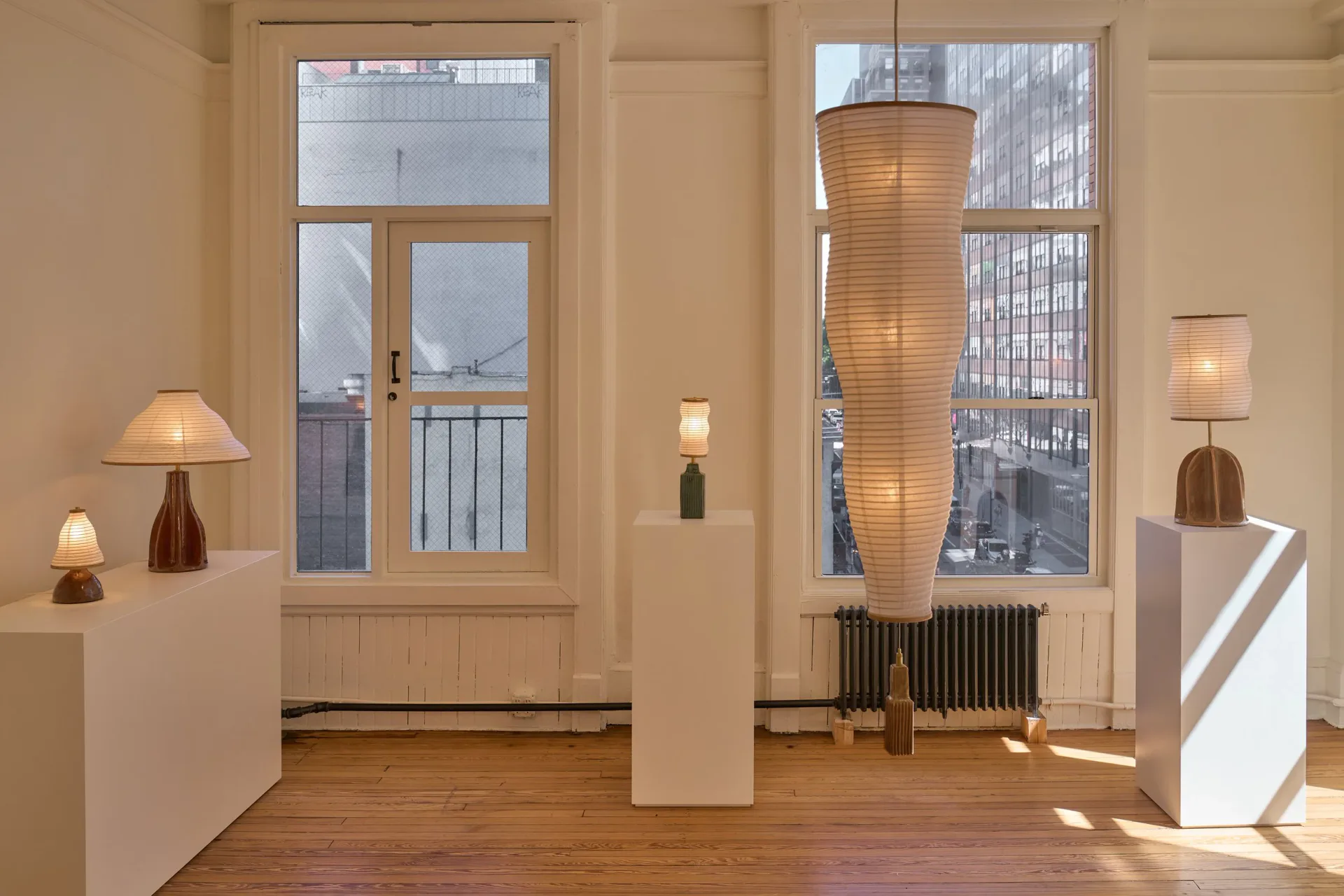 Minimalist gallery interior with wooden floors, cream walls, and tall windows; sculptural lighting installations and pedestals display work by Christopher Merchant.