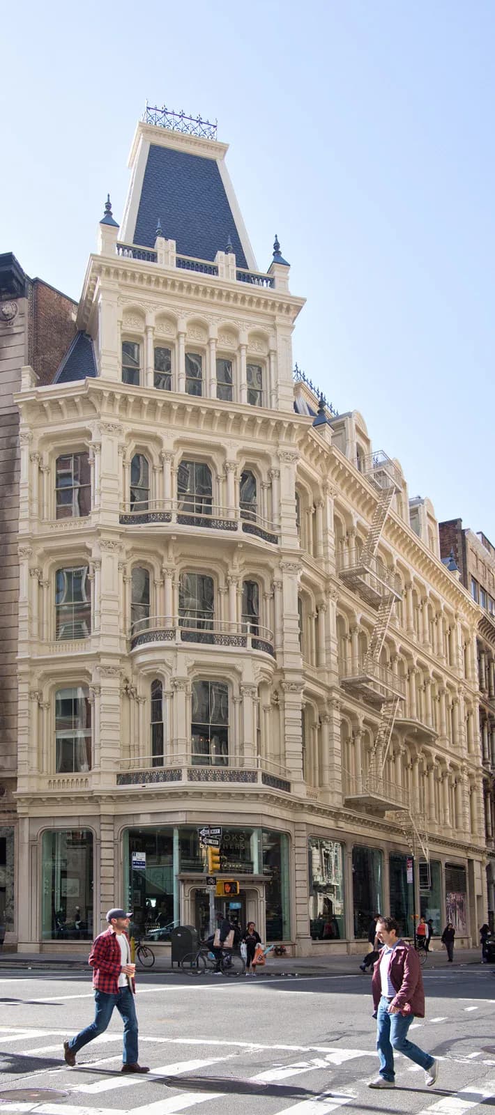 Ornate Beaux-Arts building facade with cream stone, arched windows, mansard roof with blue-slate detail, and ground-level retail, designed by Christopher Scholz.