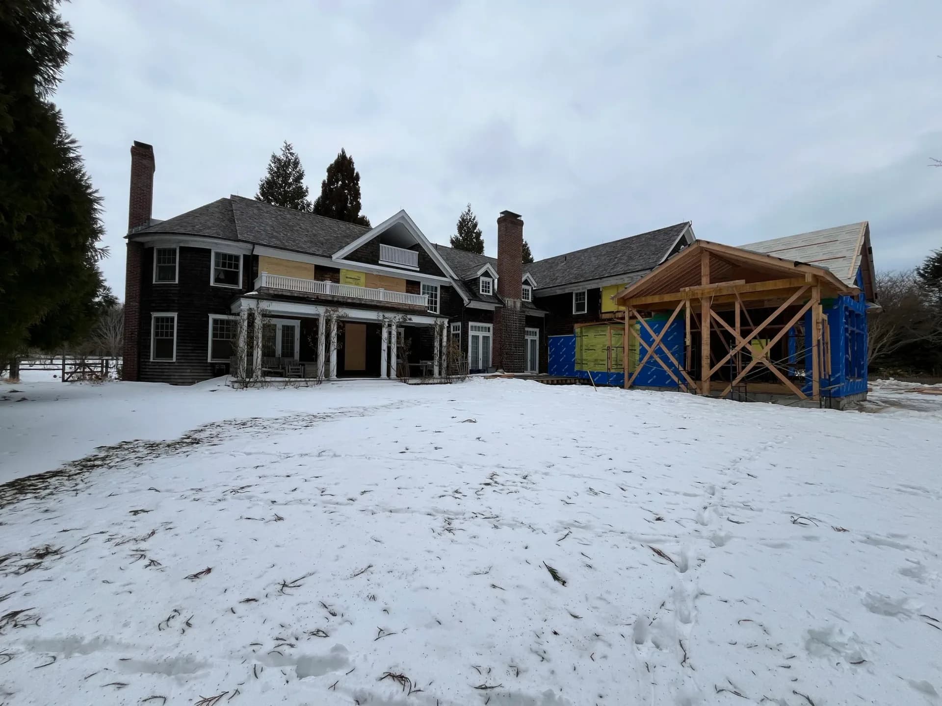 Large colonial-style residential estate with dark exterior, white trim, and an attached yellow-framed construction structure in snowy landscape by Christopher Sondi.
