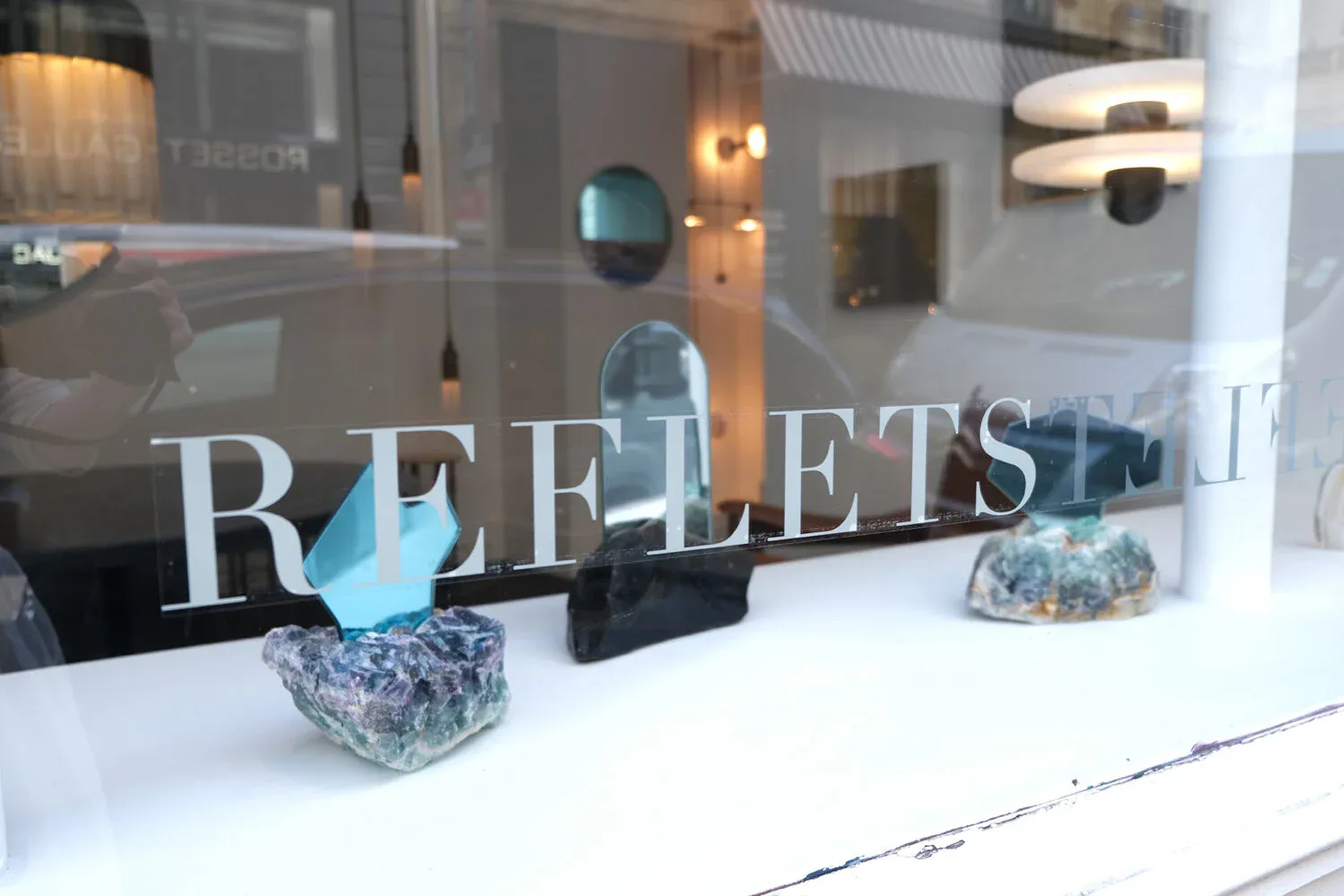 Jewelers storefront window displaying 'REFLETS' signage with blue and crystalline gemstone displays against warm interior lighting by Christopher Stuart.