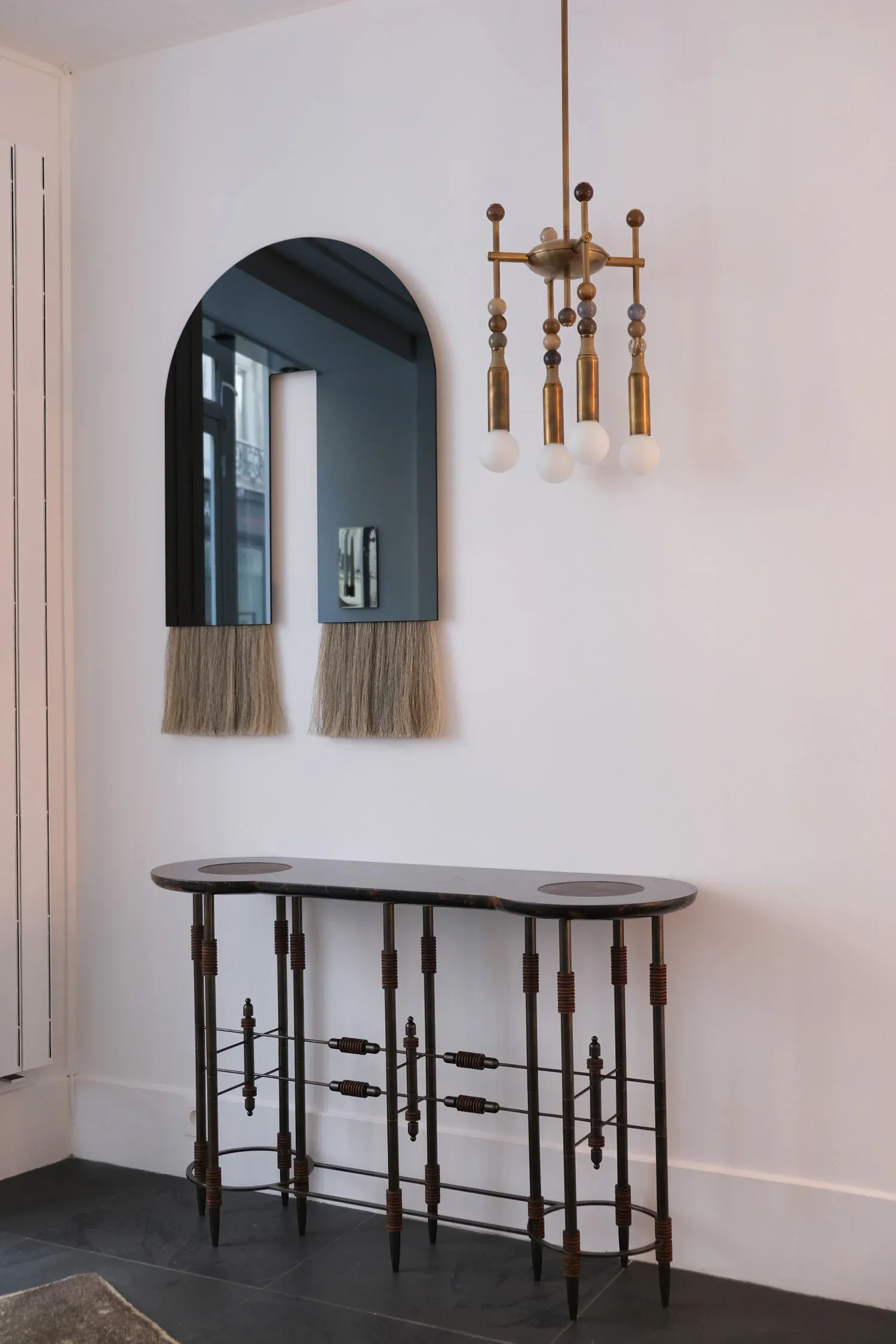 Minimalist entryway featuring a black arched mirror with natural fiber trim, bronze sculptural chandelier, and dark metal console table by Christopher Stuart.
