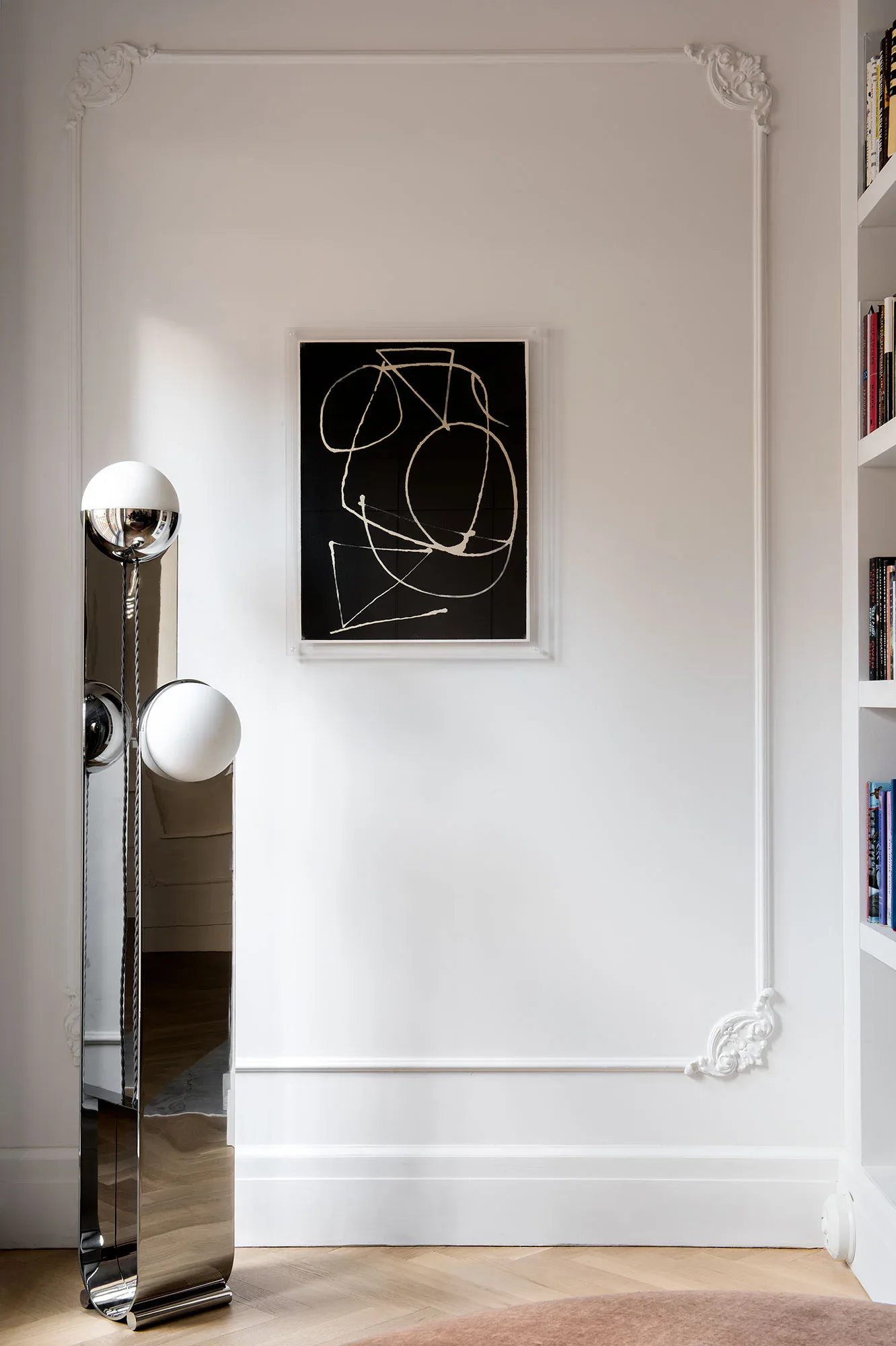 Minimalist interior with white walls, ornamental plaster corners, abstract black artwork with gold lines, polished floor lamp, and built-in shelving by Cindy Coscoros.