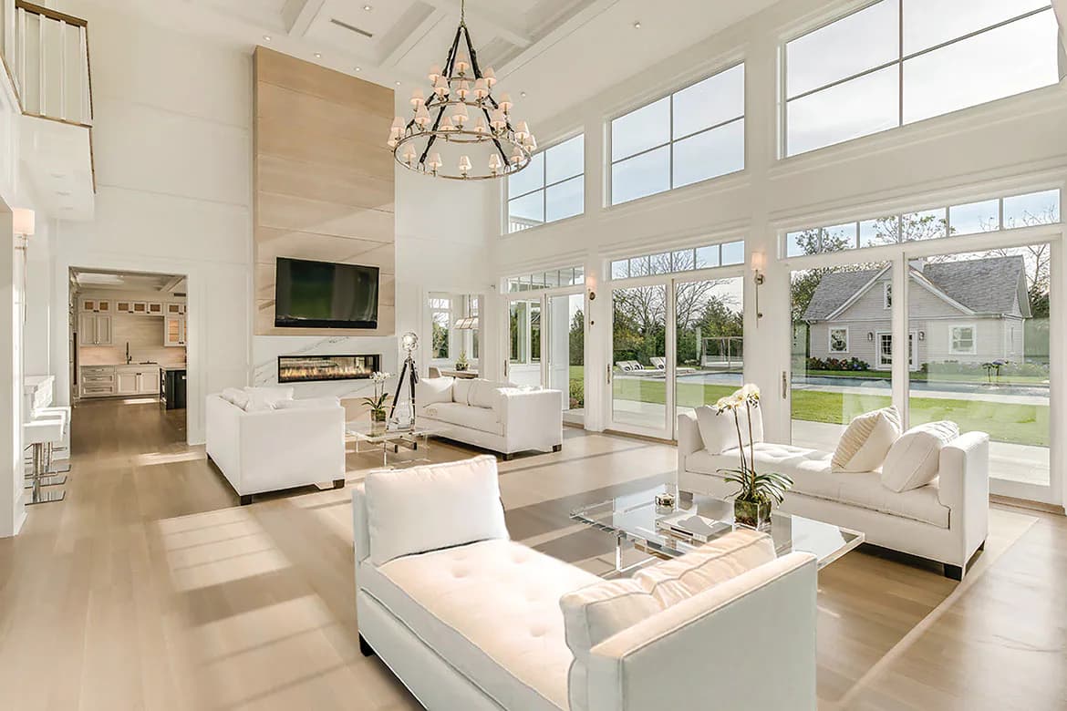 Bright, minimalist living room with white furniture, wood flooring, high ceilings, and extensive windows overlooking green lawn, designed by Cindy Montgomery.