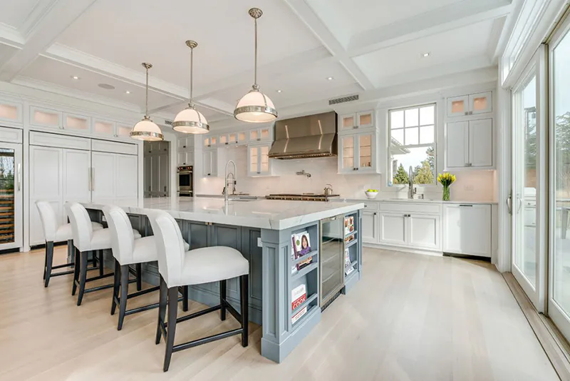 Modern kitchen with white cabinetry, gray island, pendant lighting, and seating area overlooking outdoor space, designed by Cindy Montgomery.